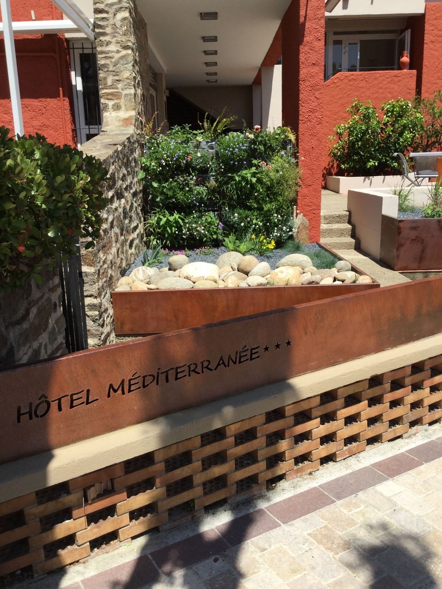 A hotel entrance with a stone wall, a planter with rocks and plants, and a sign that reads "Hôtel Méditerranée".