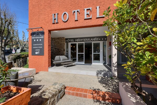 Entrada de hotel con un cartel que dice "HOTEL MÉDITERRANÉE" y servicios indicados como "CLIMATISATION", "GARAGE FERME" y "JARDIN".