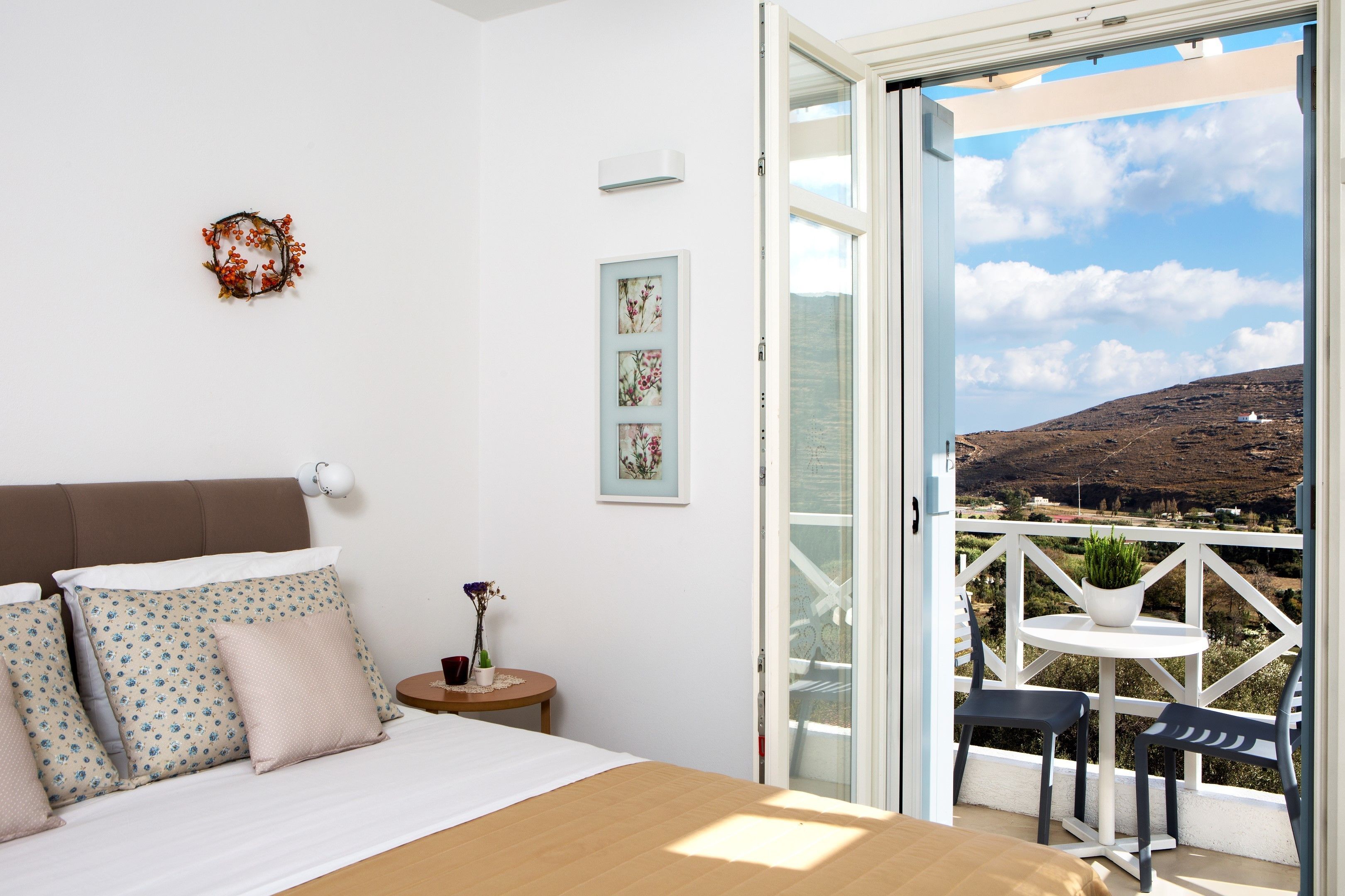 Un dormitorio con cama, almohadas, una mesita de noche, una pequeña mesa con una planta y un balcón con vistas al paisaje.