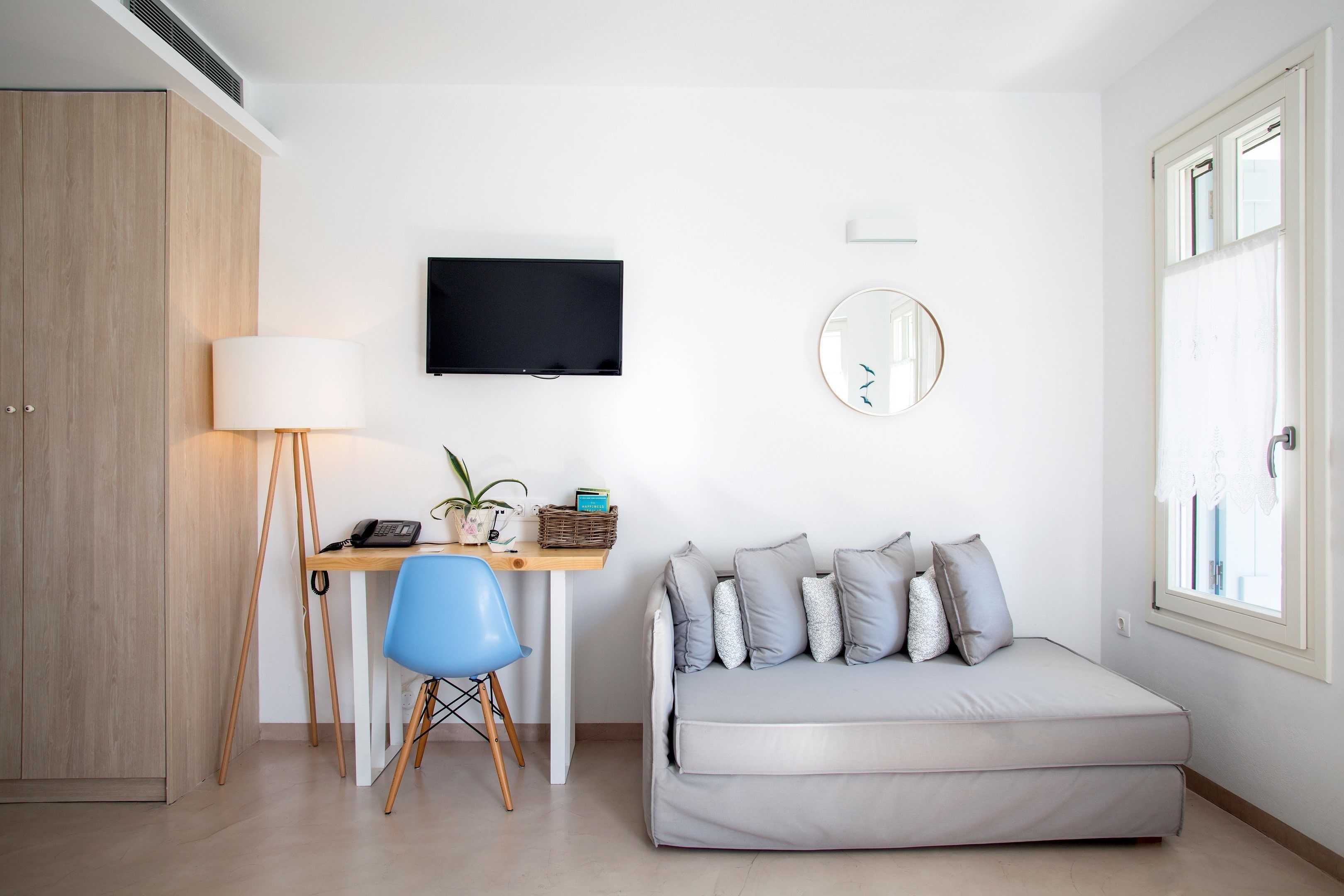 A modern, well-lit room with a wooden wardrobe, a desk with a lamp, a blue chair, a grey sofa, a flat-screen TV, and a round mirror on the wall.