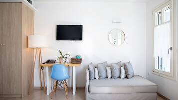 A modern, well-lit room with a wooden wardrobe, a desk with a lamp, a blue chair, a grey sofa, a flat-screen TV, and a round mirror on the wall.