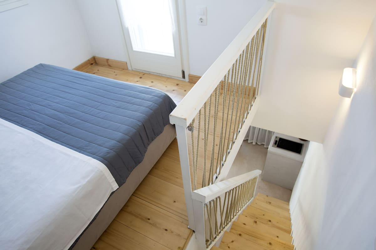 Quarto com cama, piso de madeira, corrimão branco e janela.