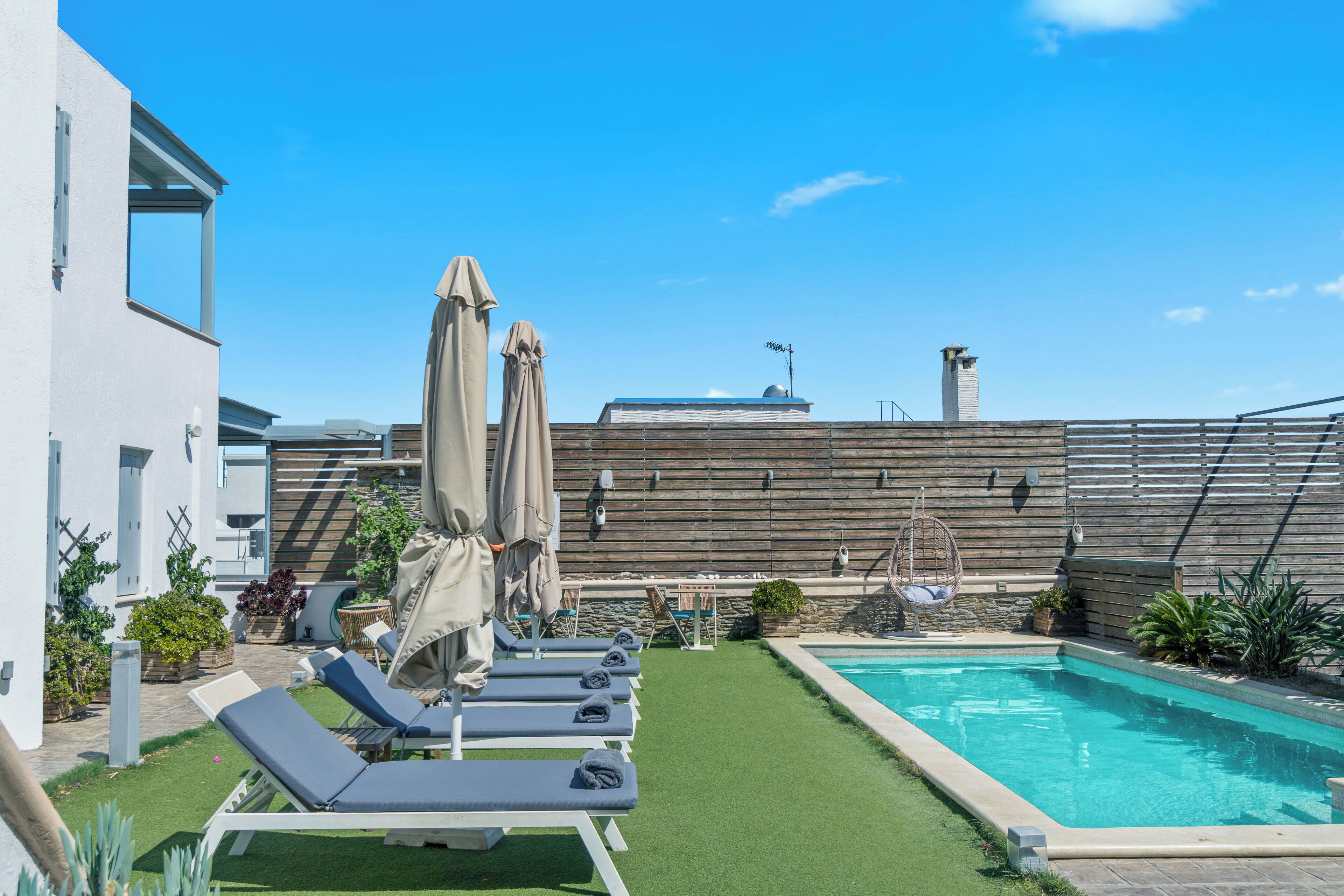 A rooftop pool area with lounge chairs and umbrellas, surrounded by a wooden fence and a building with a sloped roof.