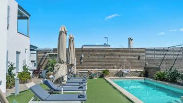 A rooftop pool area with lounge chairs and umbrellas, surrounded by a wooden fence and a building with a sloped roof.
