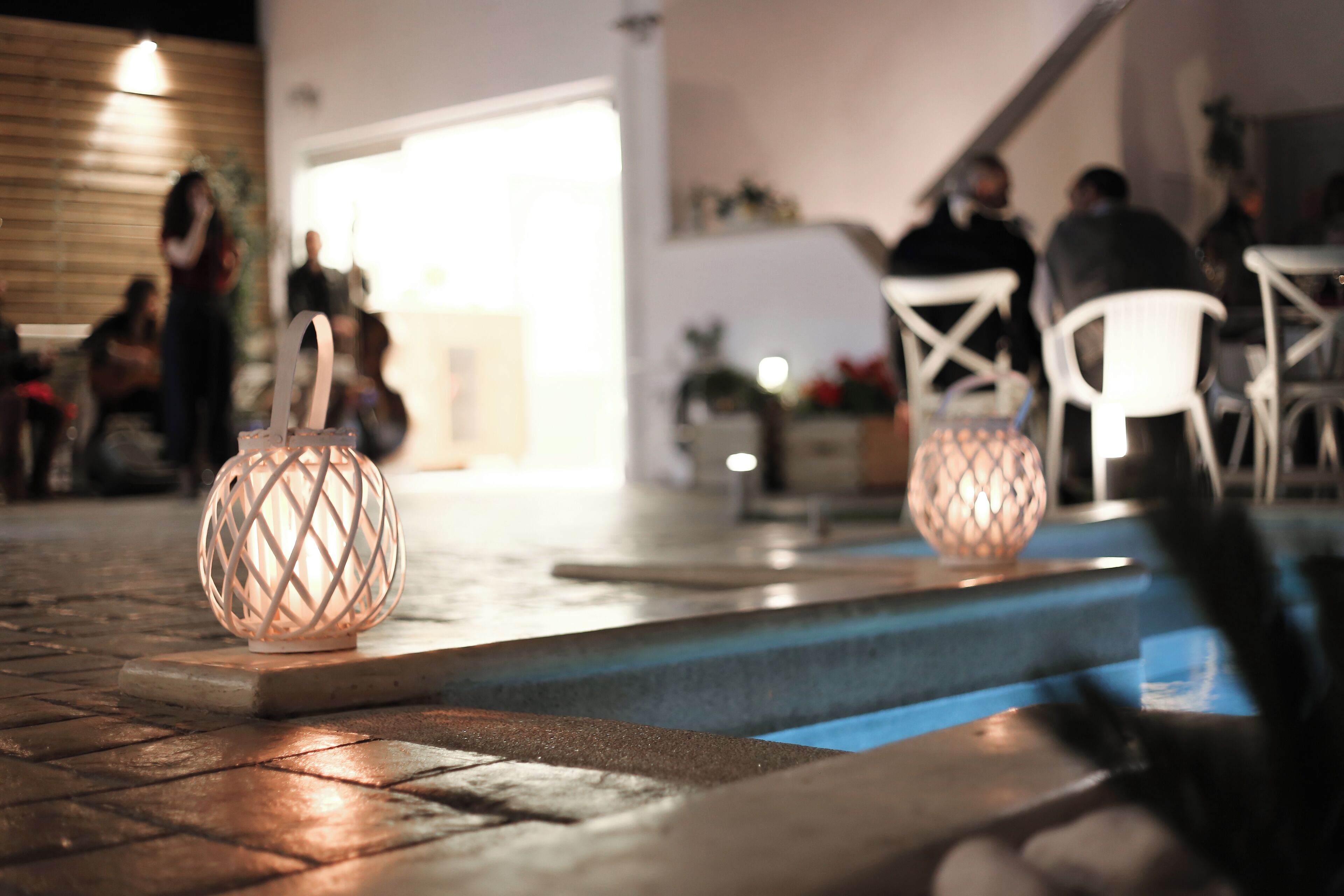 Zona de piscina con dos faroles decorativos iluminados, suelo de madera y sillas blancas.