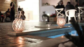 Zona de piscina con dos faroles decorativos iluminados, suelo de madera y sillas blancas.