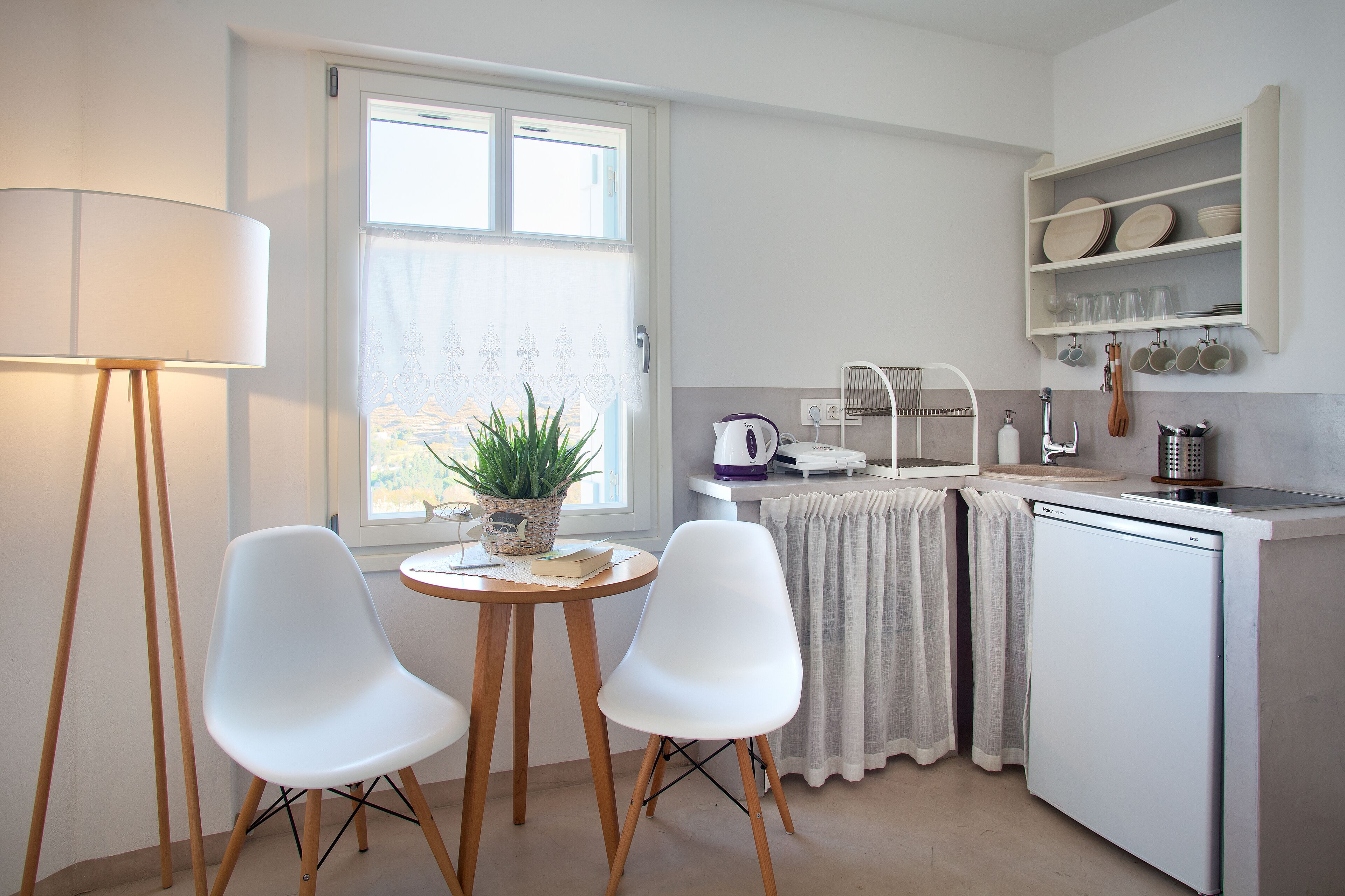 A compact kitchen with a dining area, including a table with chairs, a lamp, a potted plant, and a refrigerator.