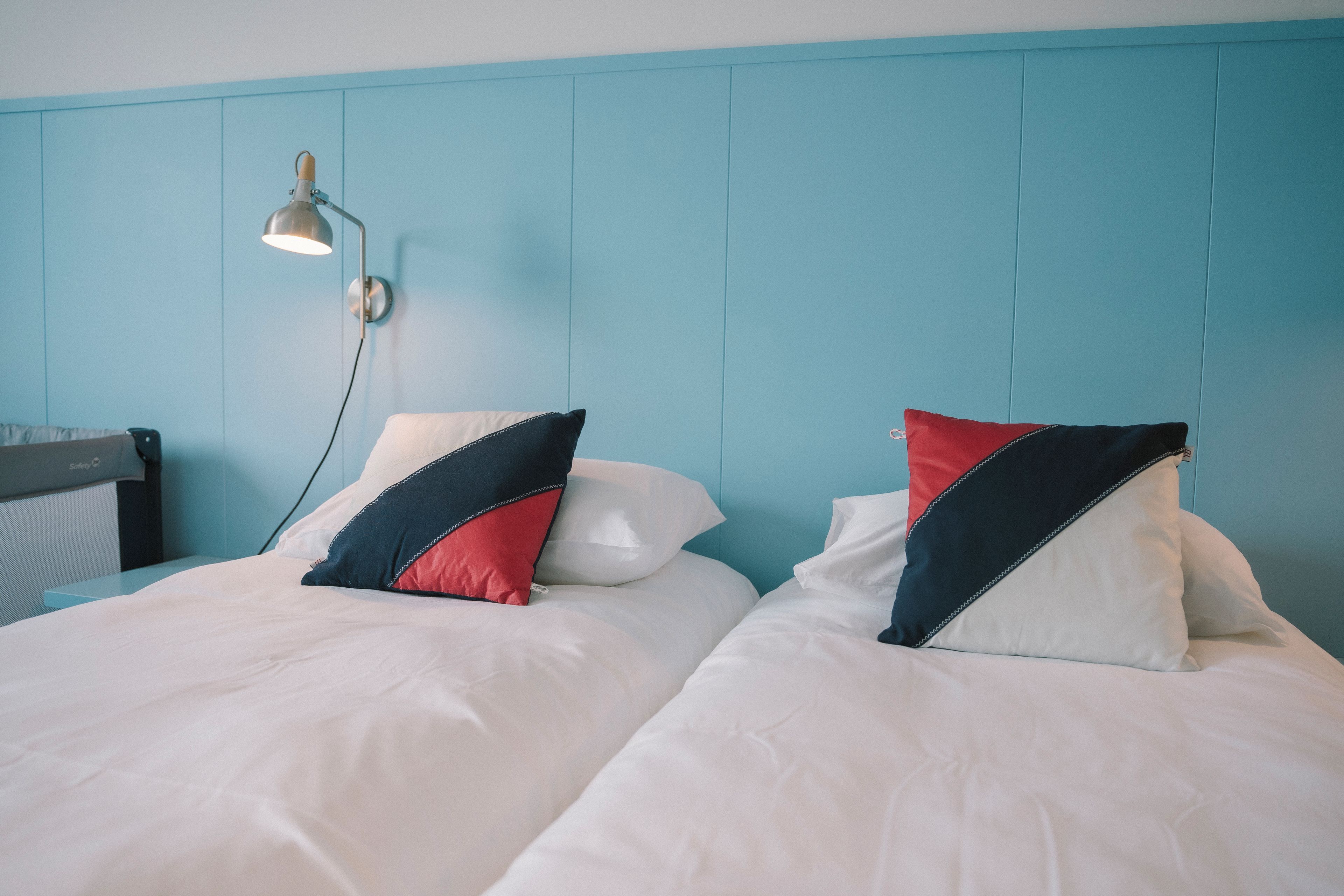 A twin bed with white bedding and two pillows, one red and one blue, in a room with a teal-colored headboard and a wall-mounted lamp.