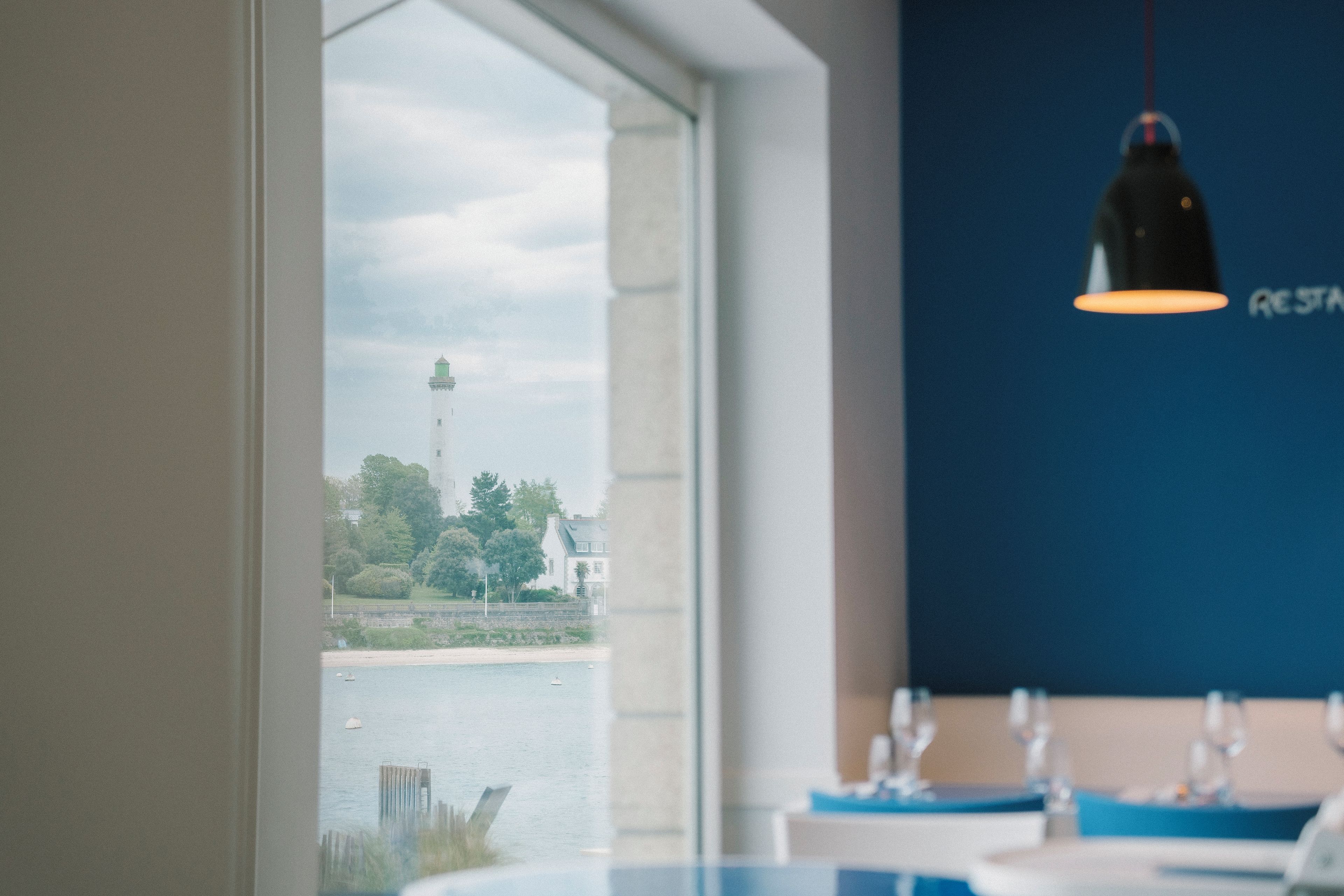 A view from a window overlooking a body of water with a lighthouse in the distance, a dining area with tables and chairs, and a hanging light fixture.