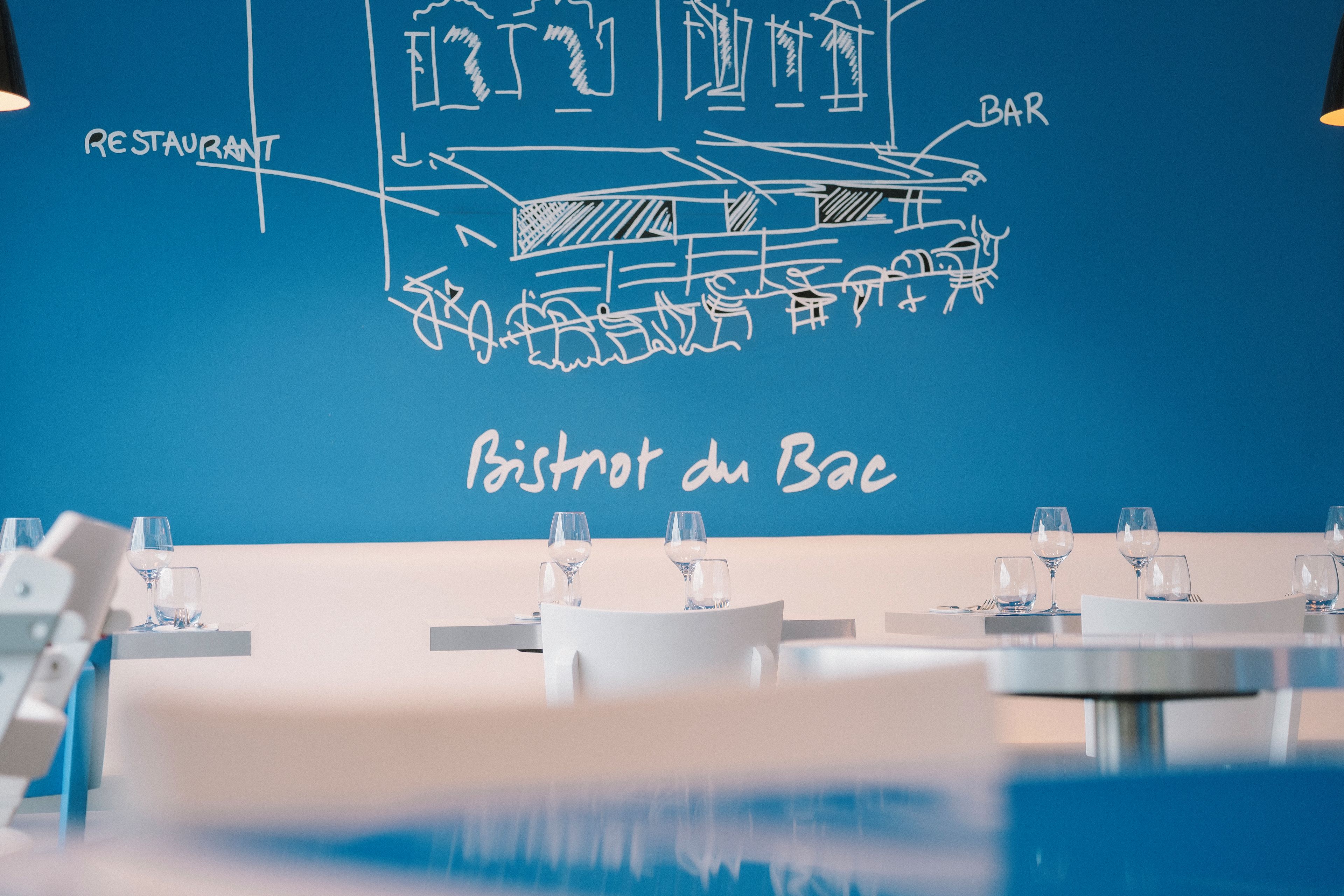 A restaurant interior with tables set for dining, a blue wall with a bar and restaurant signage, and a chalkboard menu.