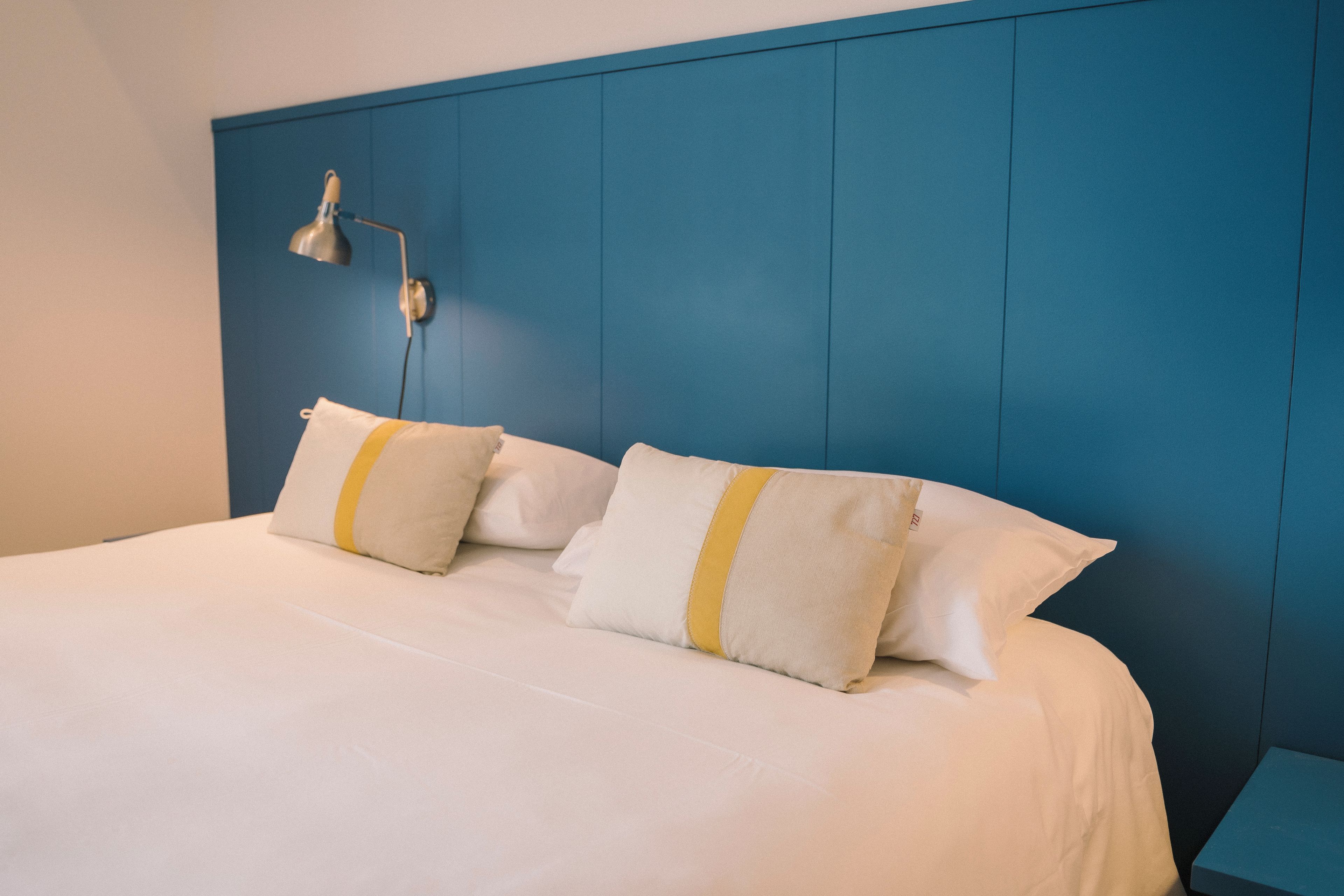 Un lit avec une literie blanche et deux oreillers, une tête de lit bleue et une table de chevet avec une lampe.