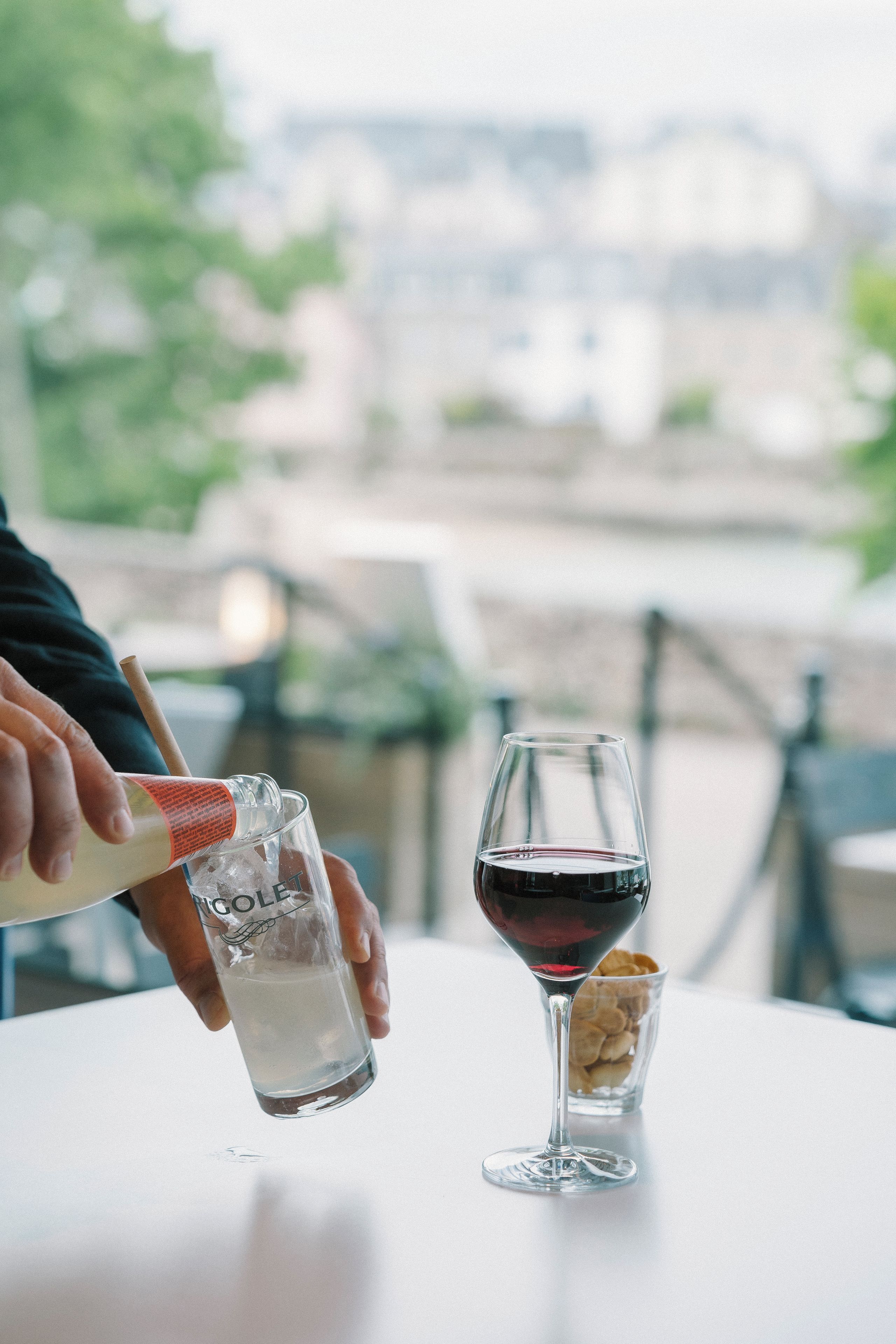 A person pouring a clear liquid into a glass with a red liquid, and a small bowl of nuts on a table.
