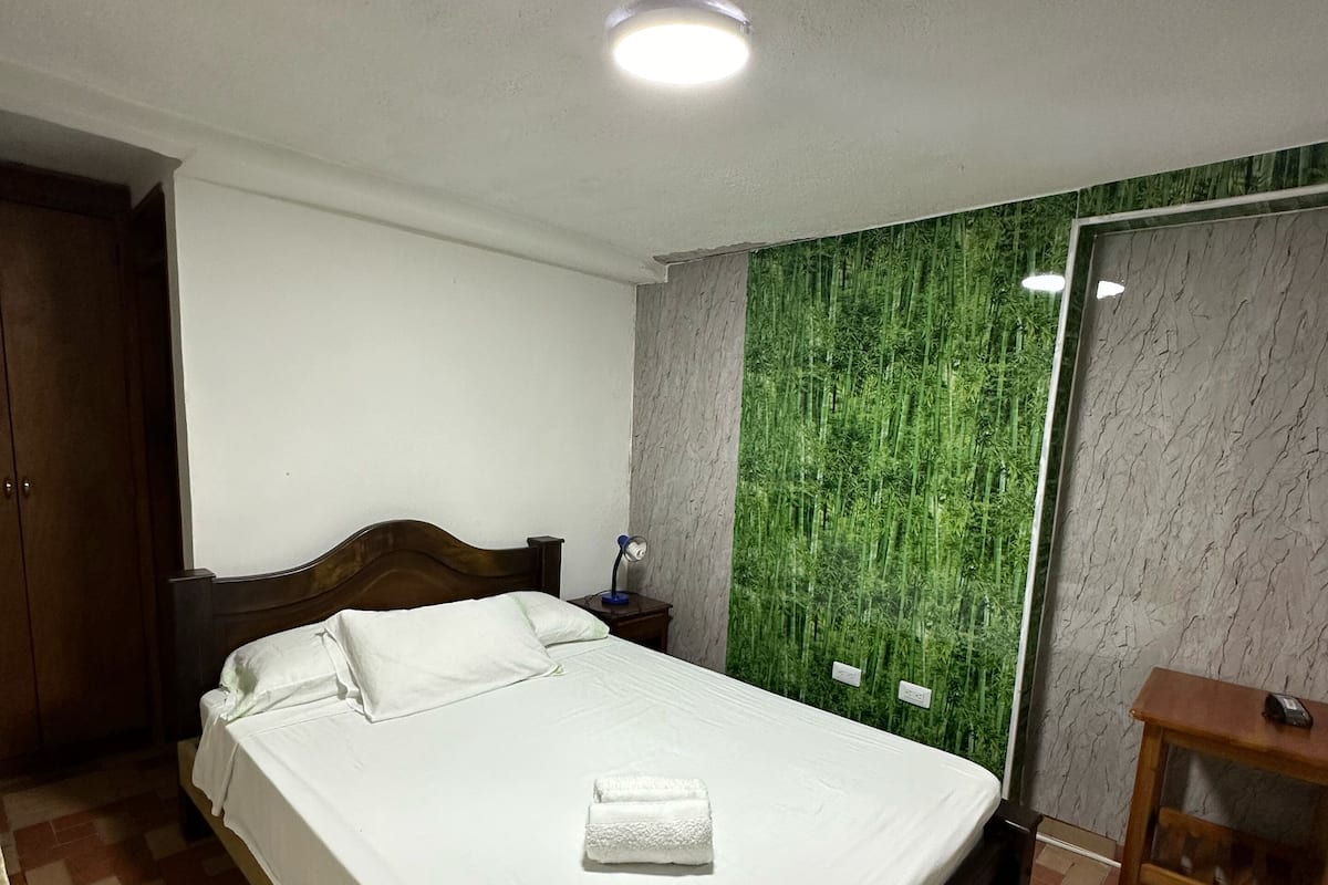 Ein ordentlich bezogenes Bett mit weißer Tagesdecke und Kissen, ein Holz-Kopfschild, ein Nachttisch mit Lampe, eine grüne Strukturwand und ein Holzschrank.