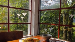 A table with two plates of food and a cup of coffee, set against a window with a view of lush greenery and bird silhouettes.