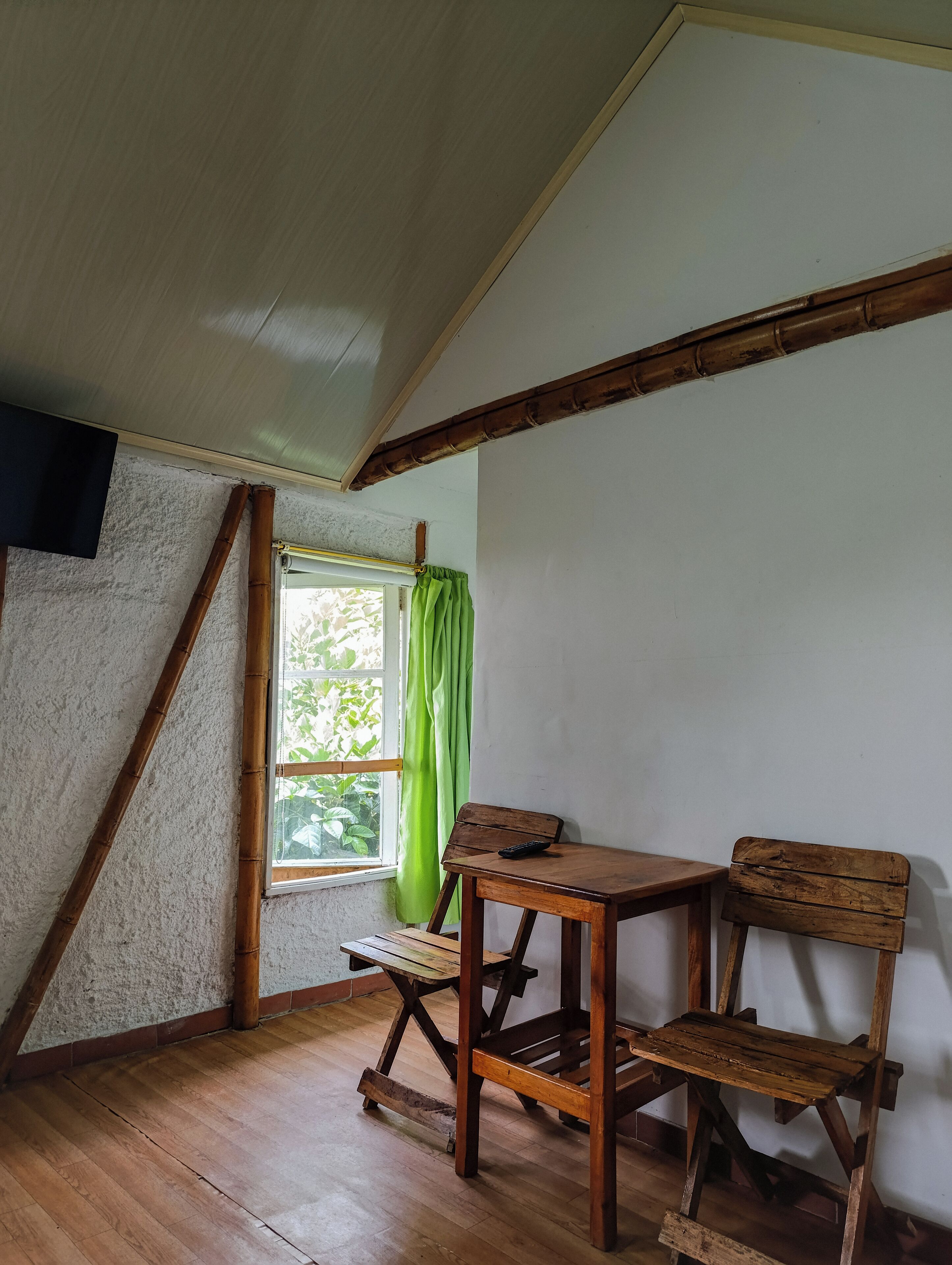 A room with a sloped ceiling, wooden beams, a window with green curtains, and a wooden desk with a chair.