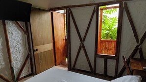 A room with a bed, two towels, a window with a view of greenery, and a wooden door.