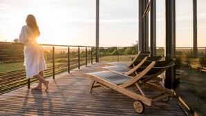 Una mujer camina sobre una terraza de madera con sillones reclinables y baranda de vidrio, con vistas a un paisaje.