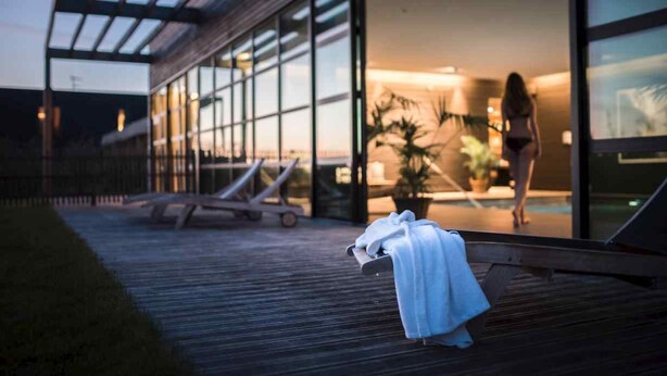 Exterior de un hotel moderno con fachada de vidrio, terraza de madera y sillones de descanso.