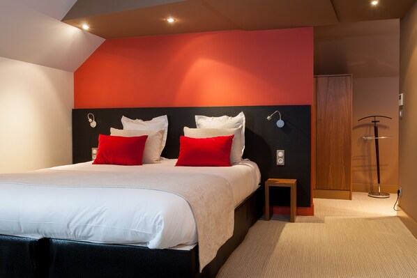 A modern hotel room with a large bed, red and white pillows, a small bedside table, and a wooden wardrobe.