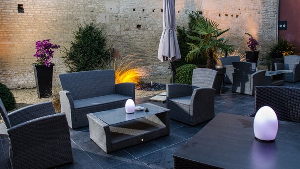 Un área de estar al aire libre con mobiliario de mimbre, una mesa con luz cálida y una pared de piedra al fondo.