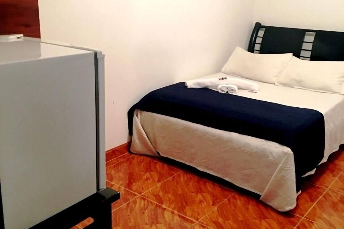 Quarto pequeno e bem organizado com uma cama de casal, uma mesa de cabeceira e uma escrivaninha.