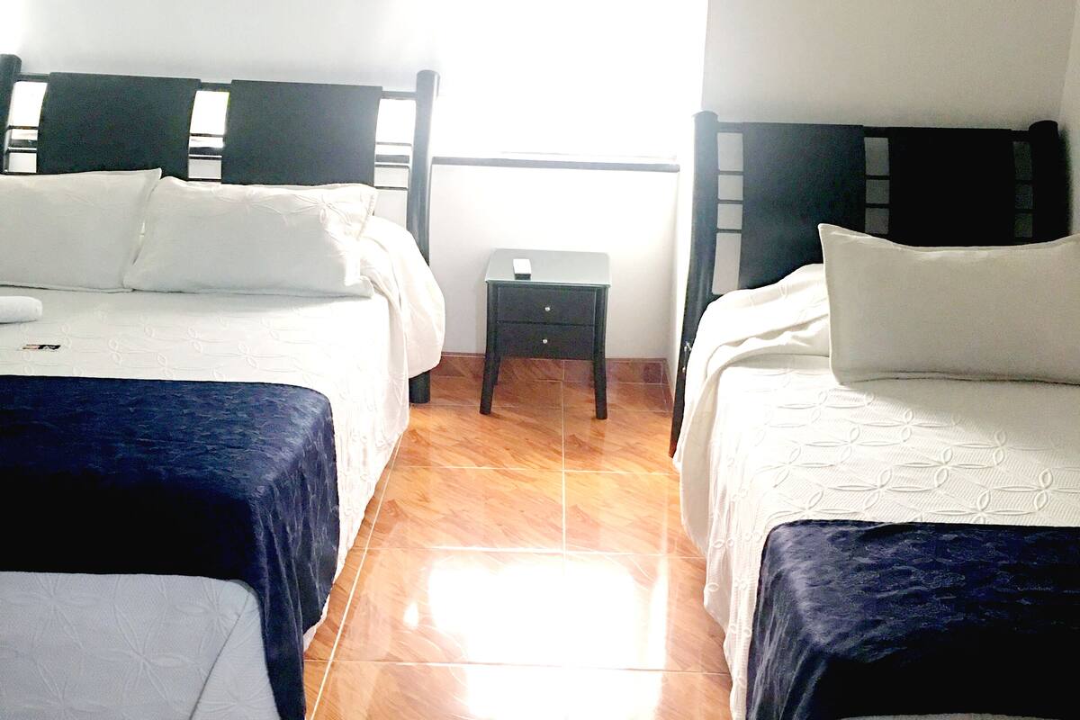 Quarto de hotel com duas camas de solteiro, uma pequena mesa de cabeceira e uma janela com persianas.