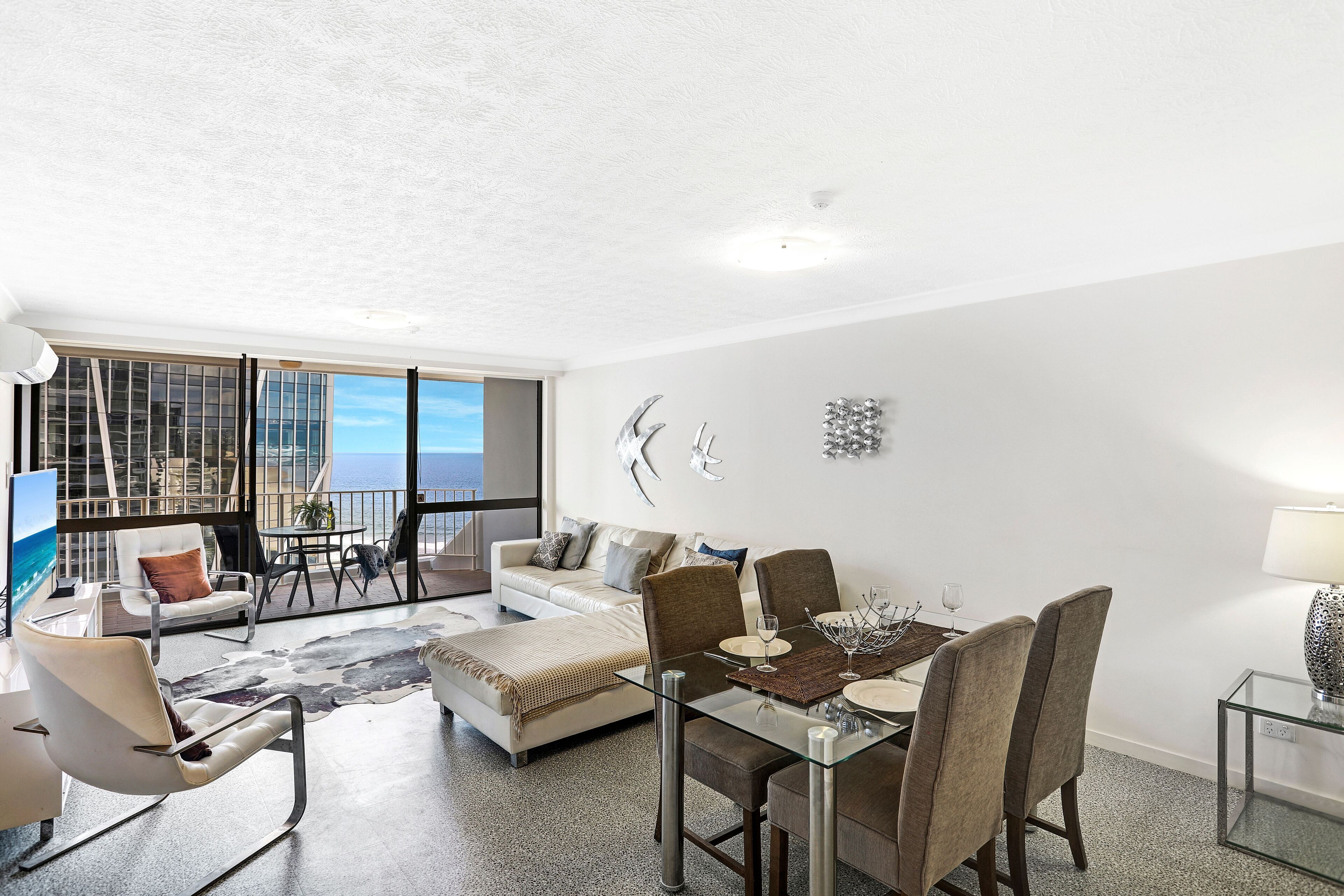 Amplia sala de estar con balcón, una mesa de comedor preparada para cuatro comensales y un gran ventanal con vista al océano.