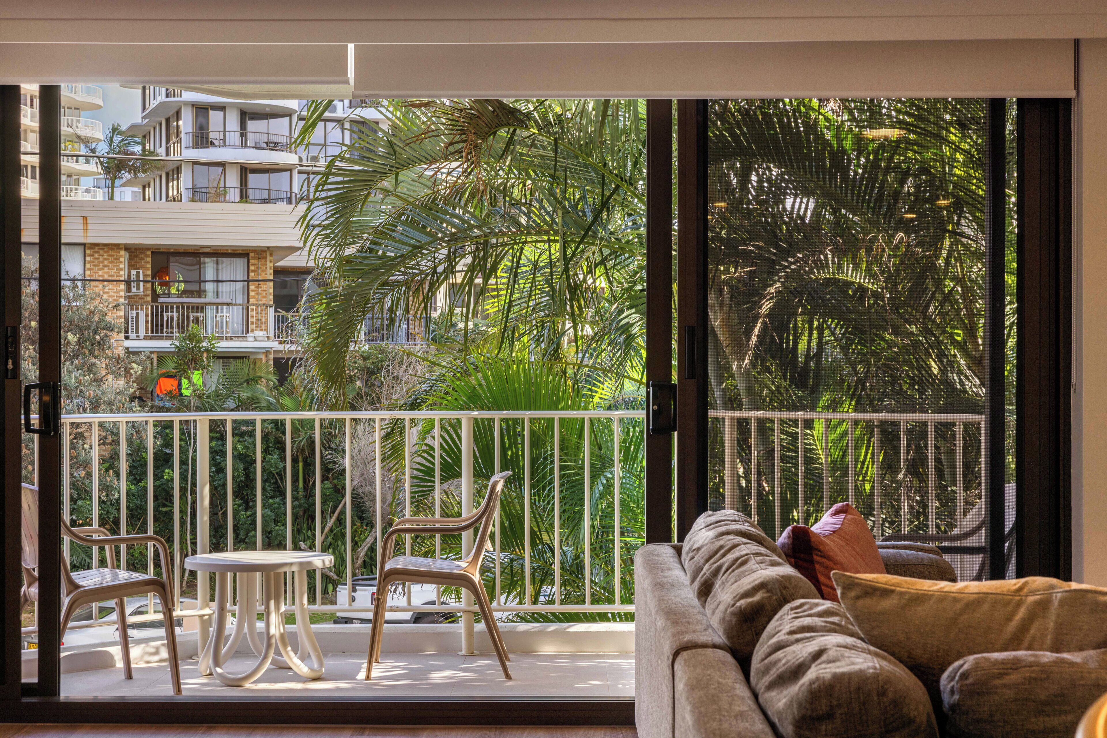 Un balcón con sofá y sillas, una mesa y vistas a un edificio y vegetación.