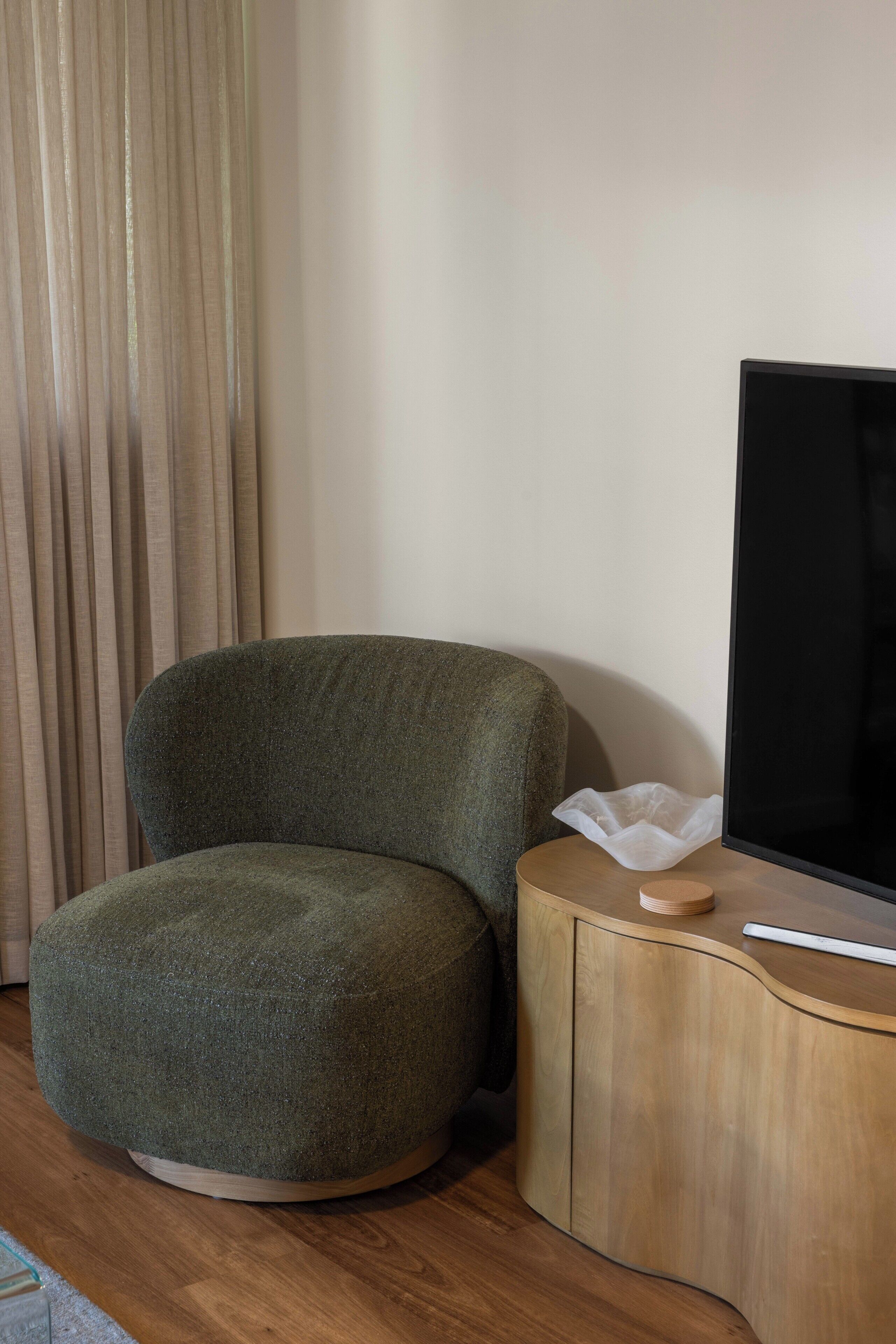 A room with a round, green upholstered chair, a wooden side table with a flat-screen TV, and a curtain.