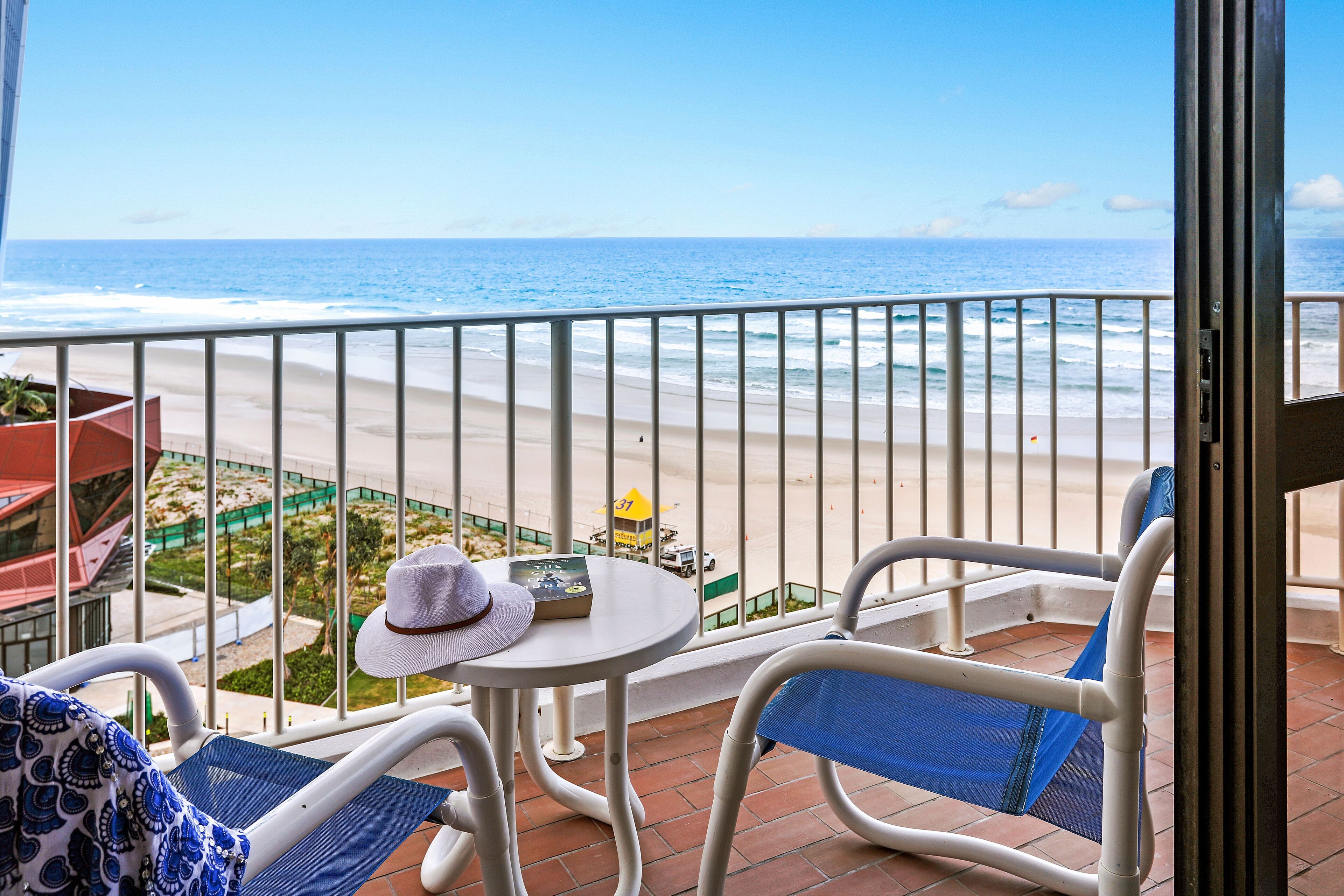 Ein Balkon mit Blick auf den Strand, ein Tisch mit Hut und Buch sowie ein Stuhl mit blauer Sitzkissen.