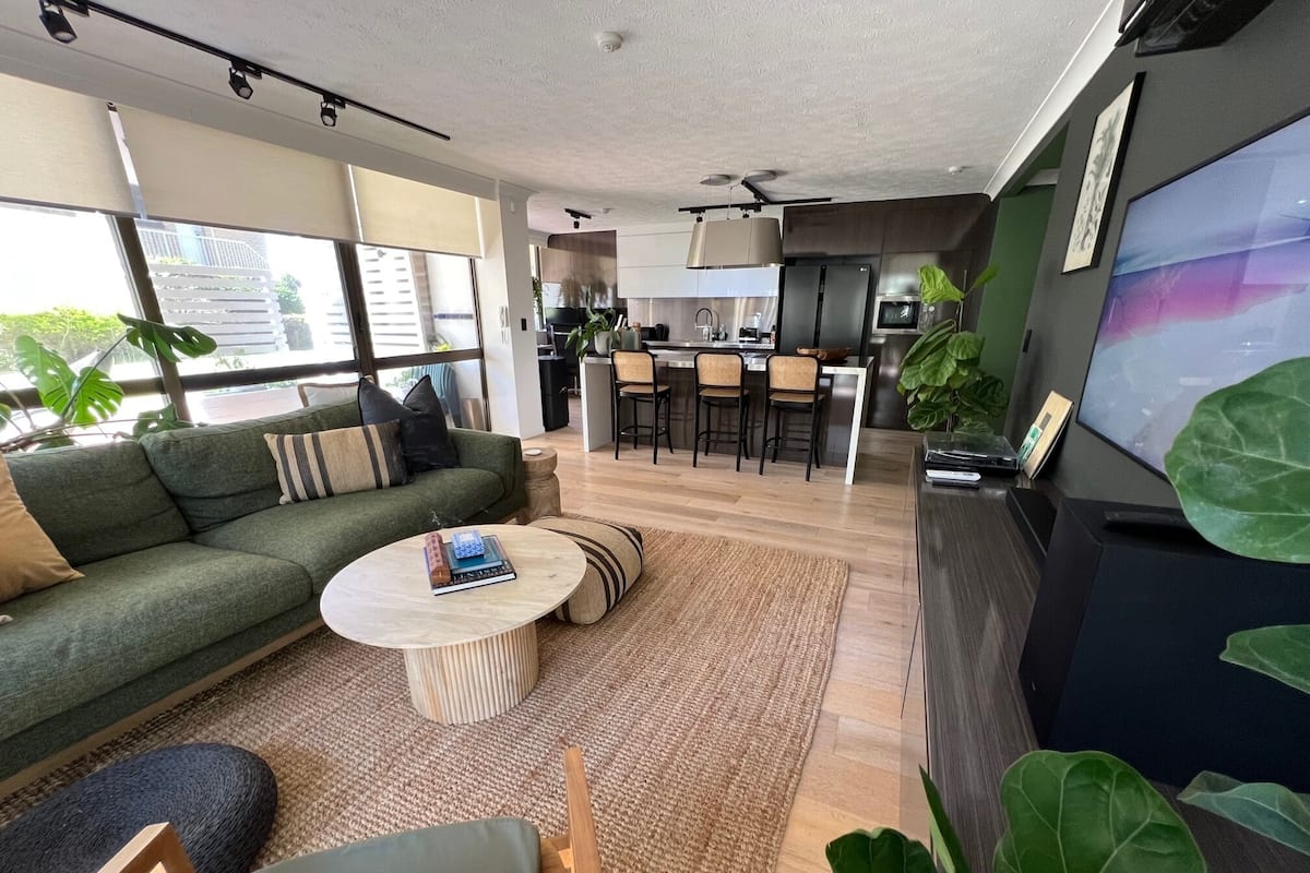 A modern living room with a sofa, coffee table, and a dining area with chairs and a kitchen in the background.