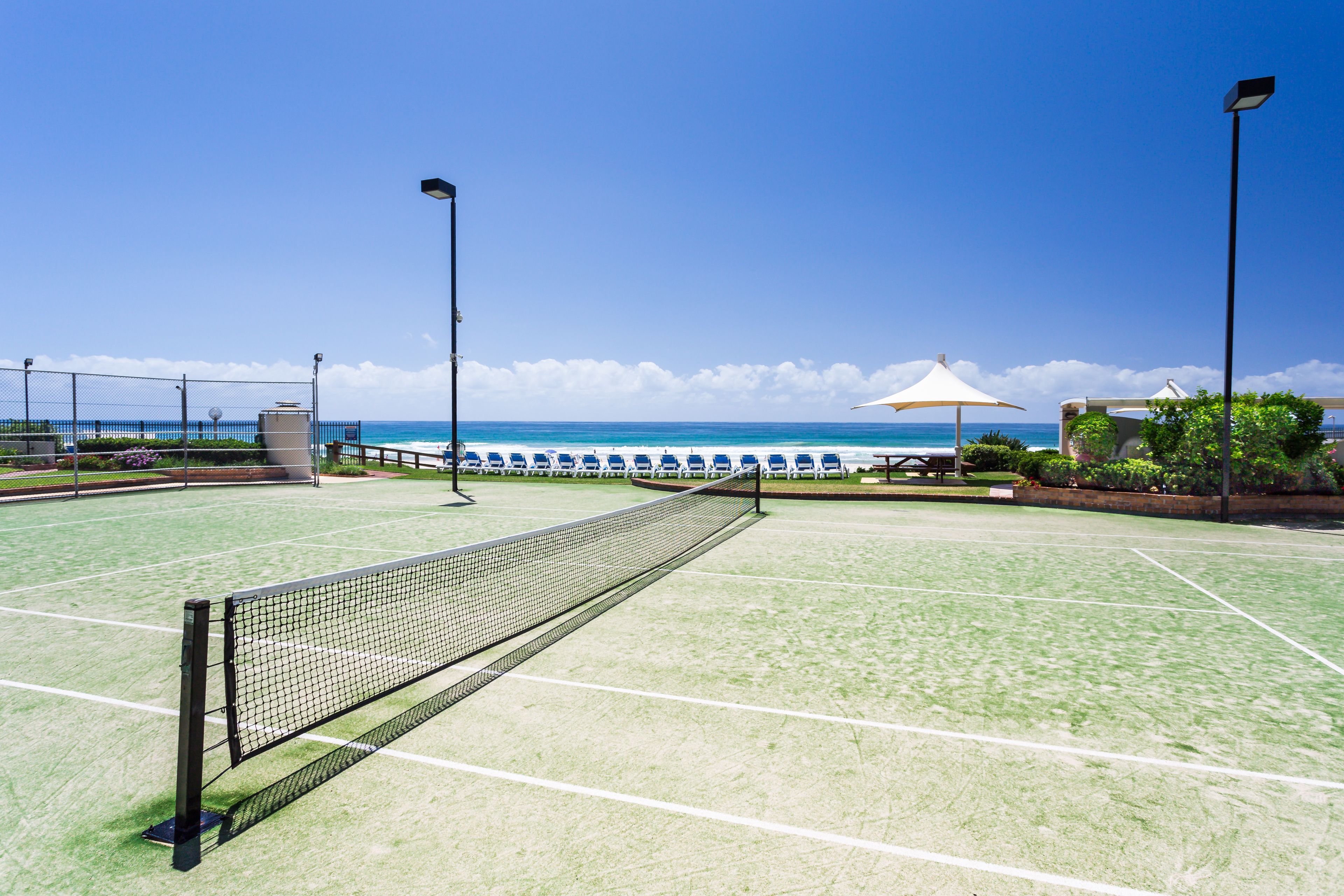 A tennis court with a net, surrounded by a fence, and a view of the ocean.
