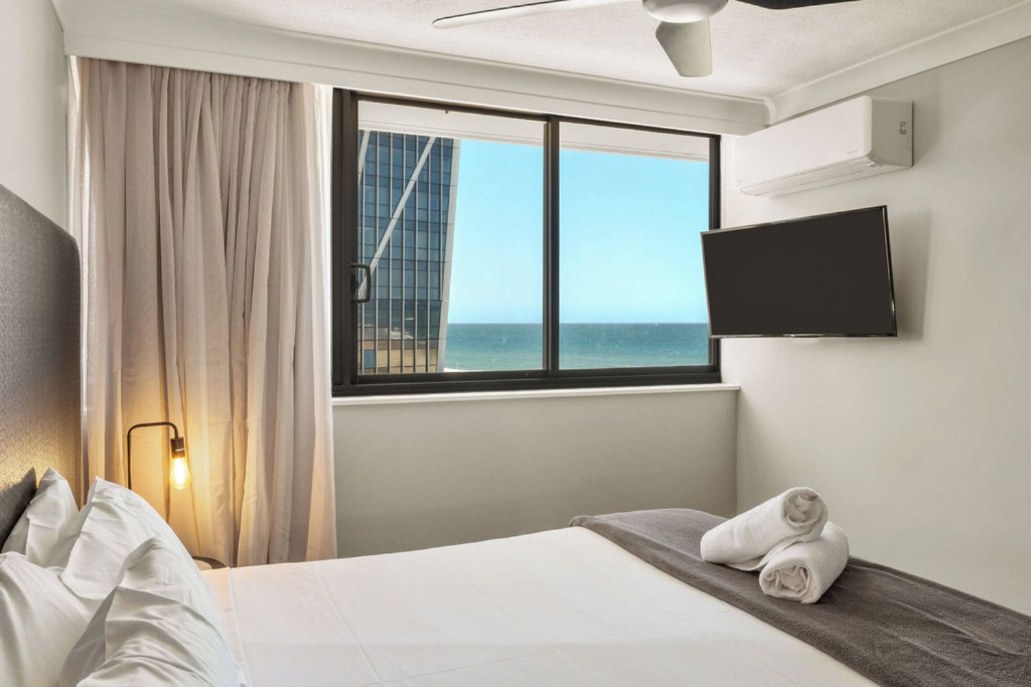 Quarto de hotel com cama, janela vista para o mar, televisão de tela plana na parede e ventilador de teto.