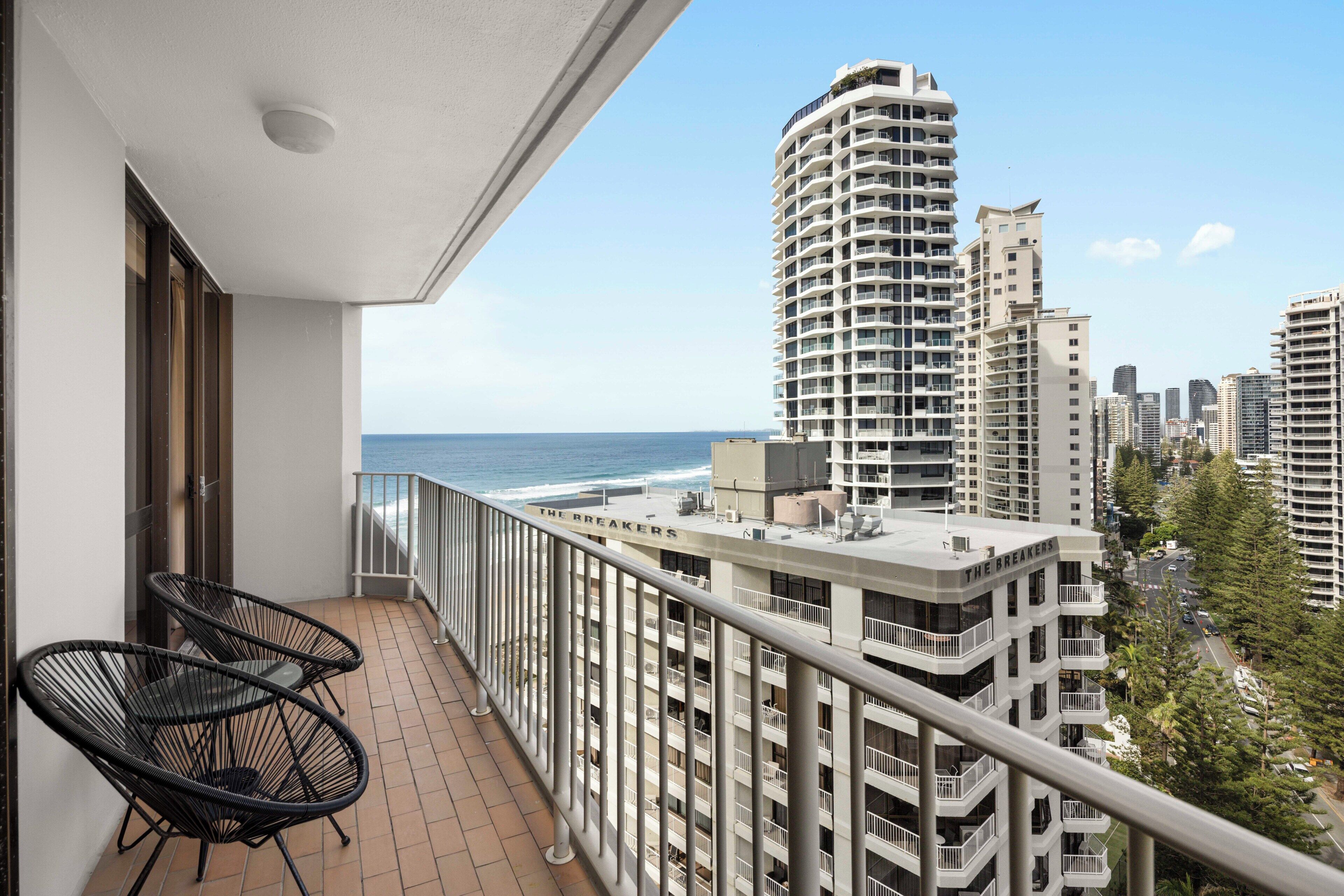 A balcony with two chairs, a view of the ocean, and high-rise buildings in the background.