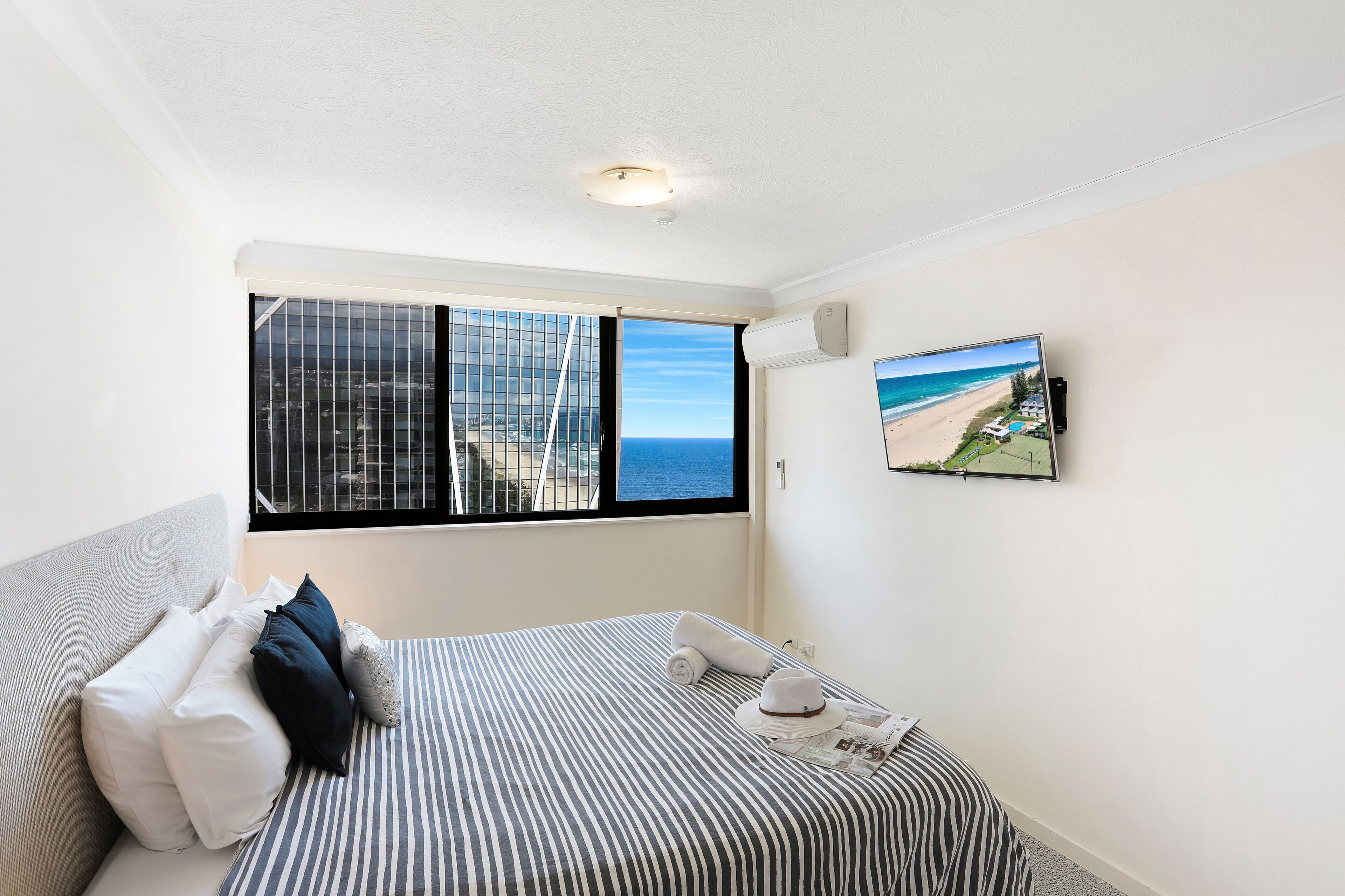 Habitación de hotel con un ventanal grande que ofrece vistas a la playa, una cama con ropa de cama a rayas y un televisor de pared que muestra una escena de playa.