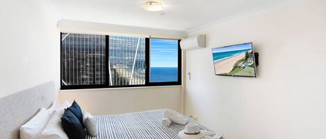 Habitación de hotel con un ventanal grande que ofrece vistas a la playa, una cama con ropa de cama a rayas y un televisor de pared que muestra una escena de playa.