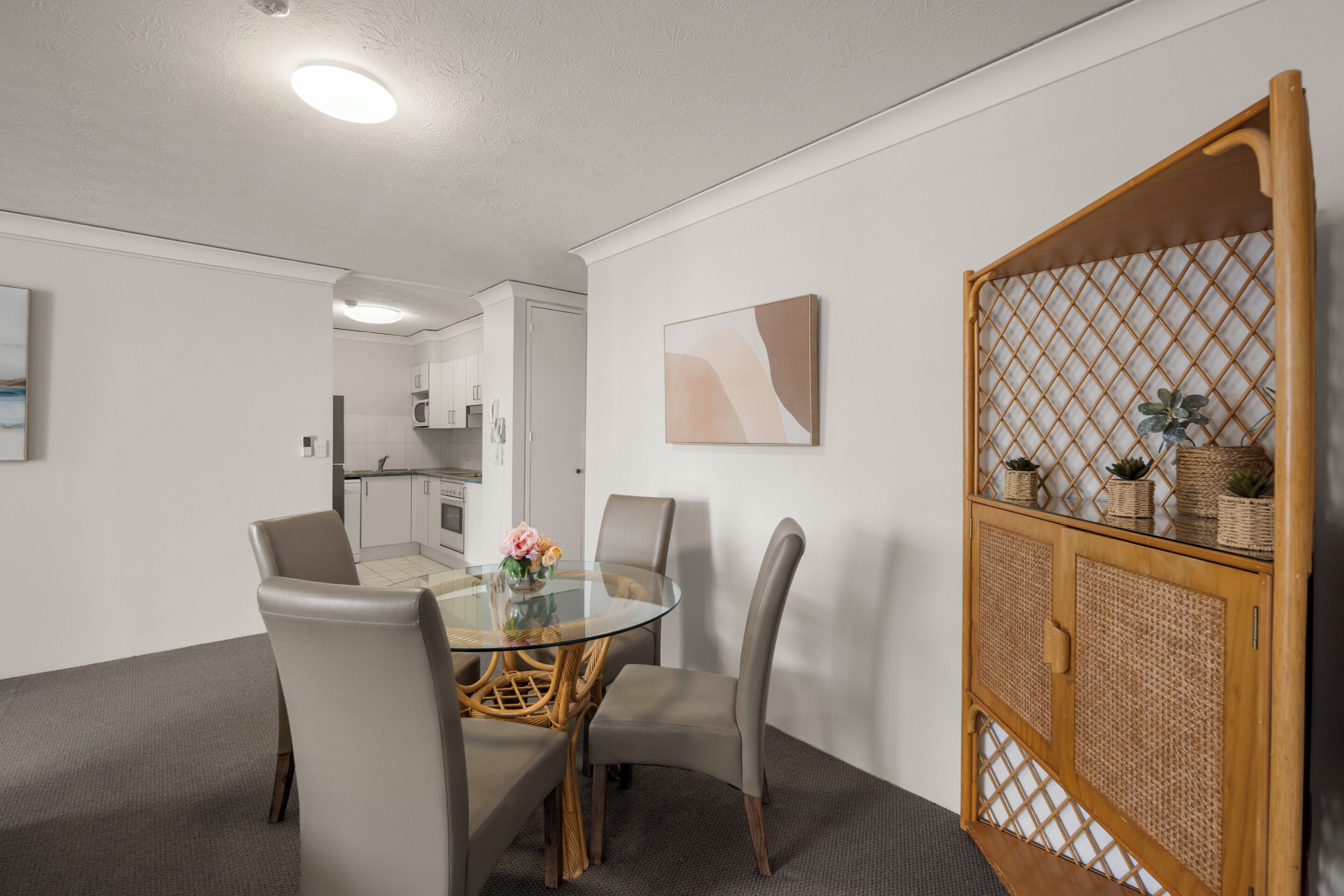 Un espace repas moderne avec une table en verre, des chaises grises, un fauteuil en osier, un meuble en bois orné de plantes et, au fond, une cuisine.