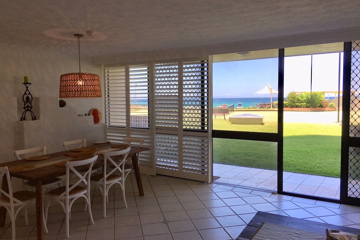 A dining area with a wooden table and white chairs, a hanging light fixture, and a view of a grassy outdoor area with a beach and a tent.