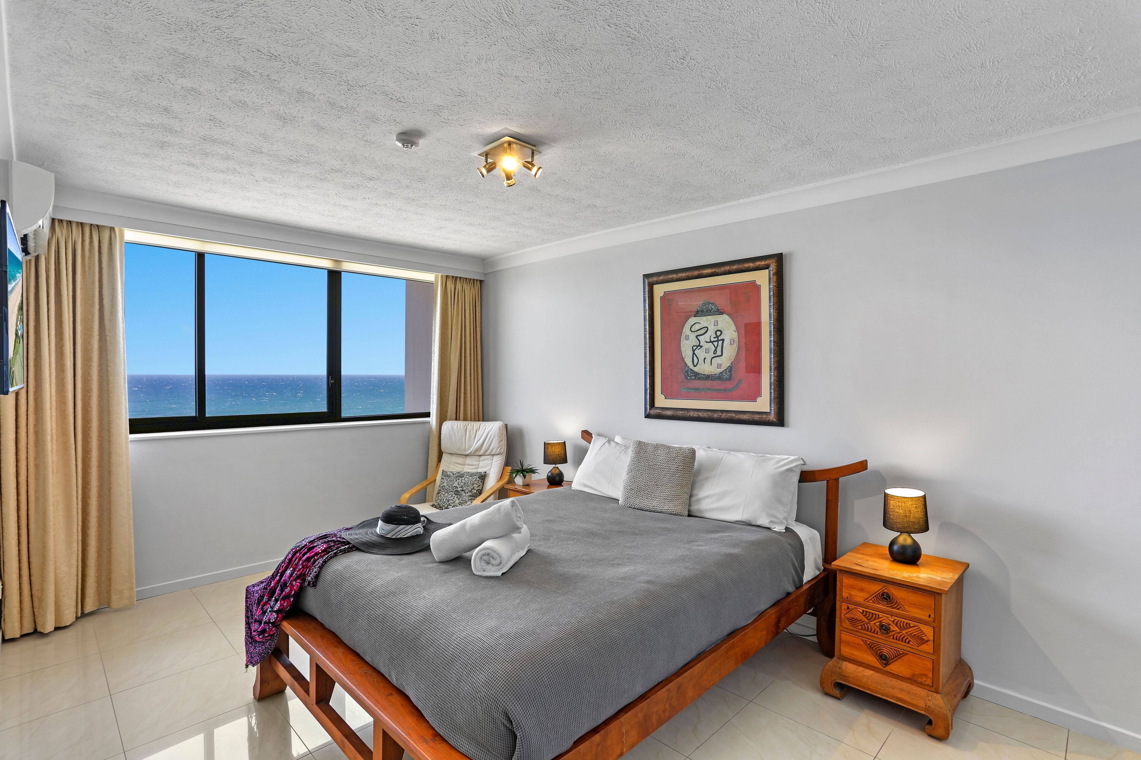 A neatly made bed with a grey blanket, a wooden nightstand with a lamp, a chair, and a large window with a view of the ocean.