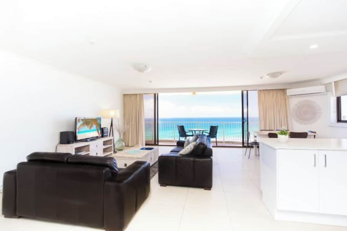 A modern living room with a large window overlooking the sea, a sofa, a coffee table, and a kitchen area.