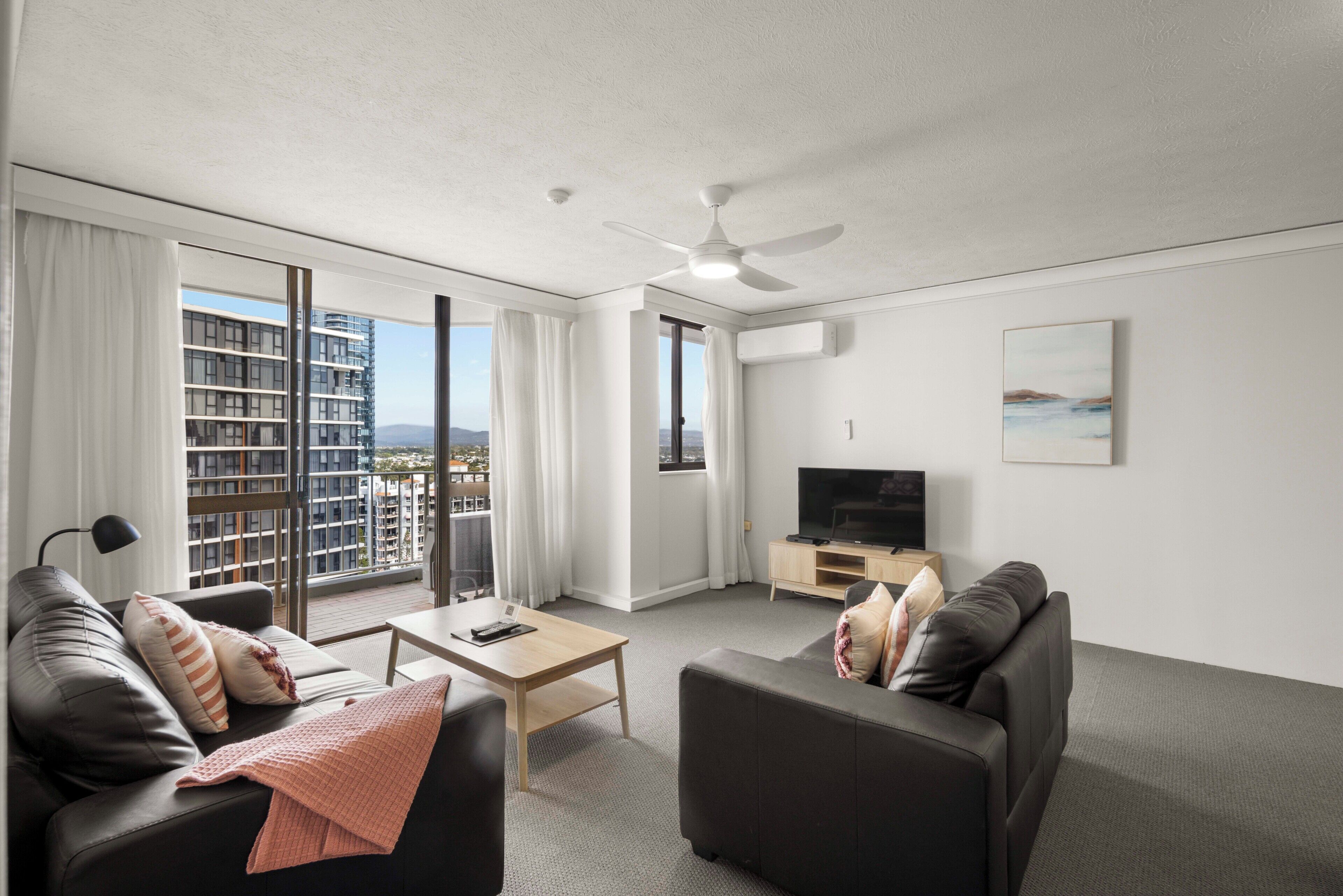 A modern hotel room with a balcony, a flat-screen TV, leather sofas, a coffee table, and a large window with curtains.