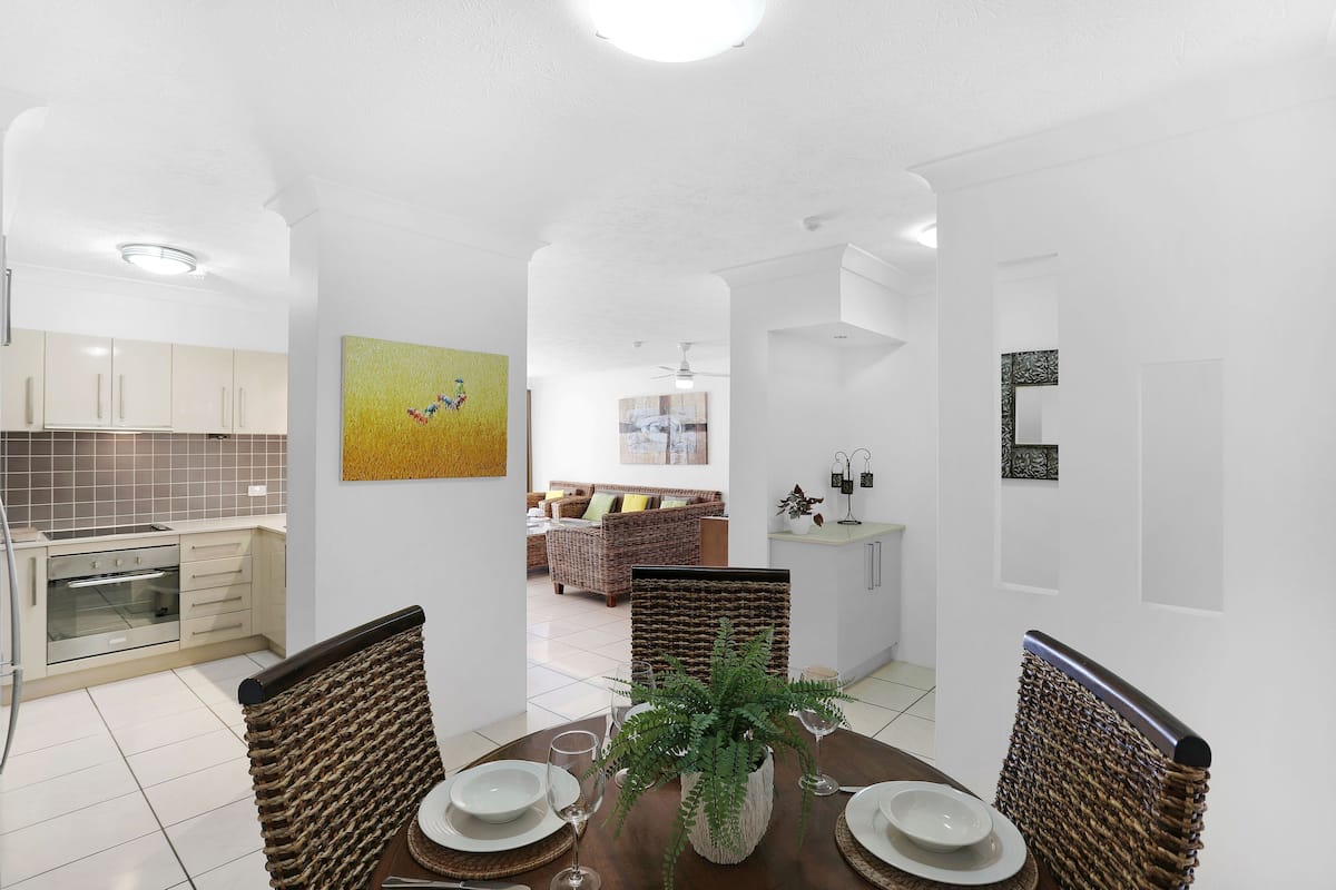 A dining area with a round table, chairs, and a central plant. A kitchen with white cabinets and a tiled backsplash is visible in the background.