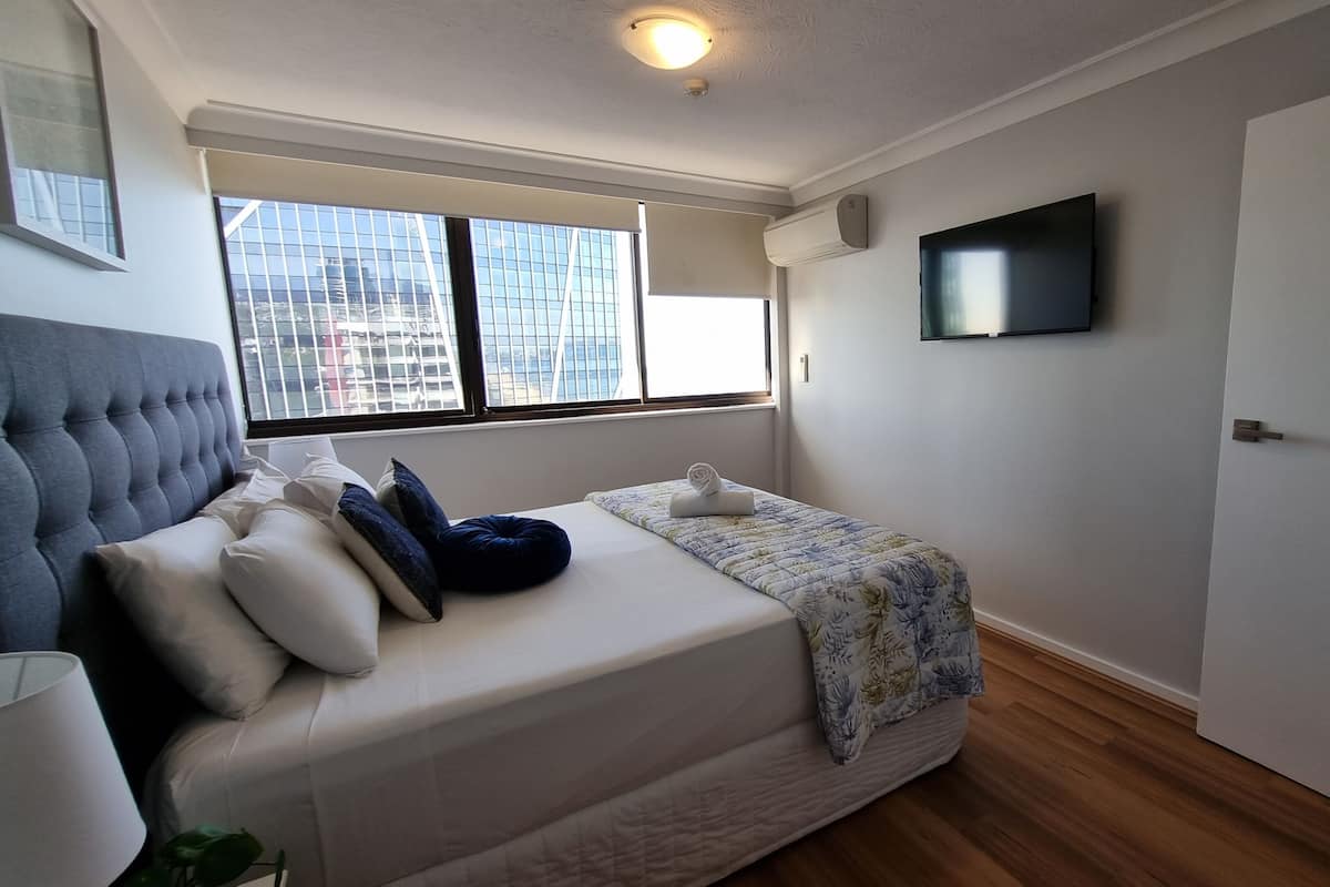 A bedroom with a bed, a TV mounted on the wall, and a window with a city view.