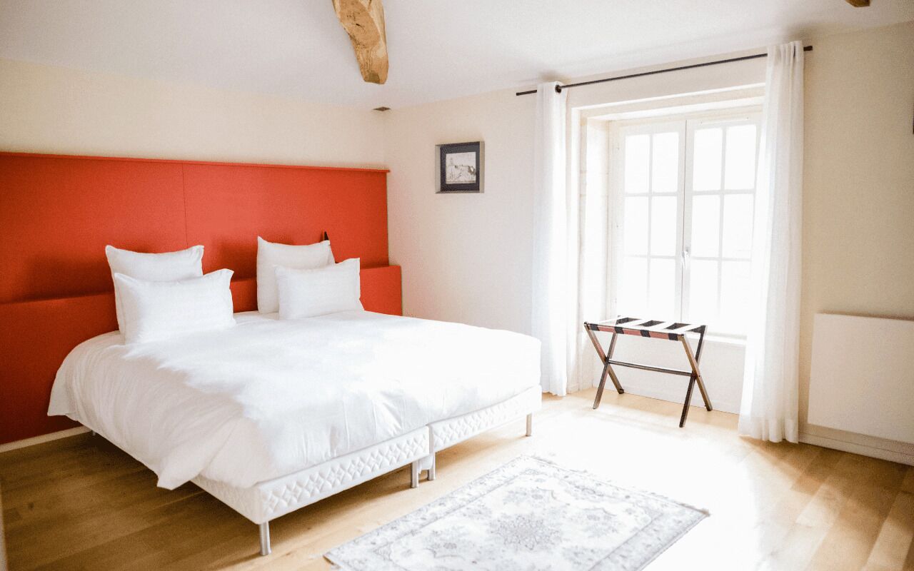 Une chambre à coucher avec un lit, une tête de lit rouge, une fenêtre avec des rideaux et une petite table.