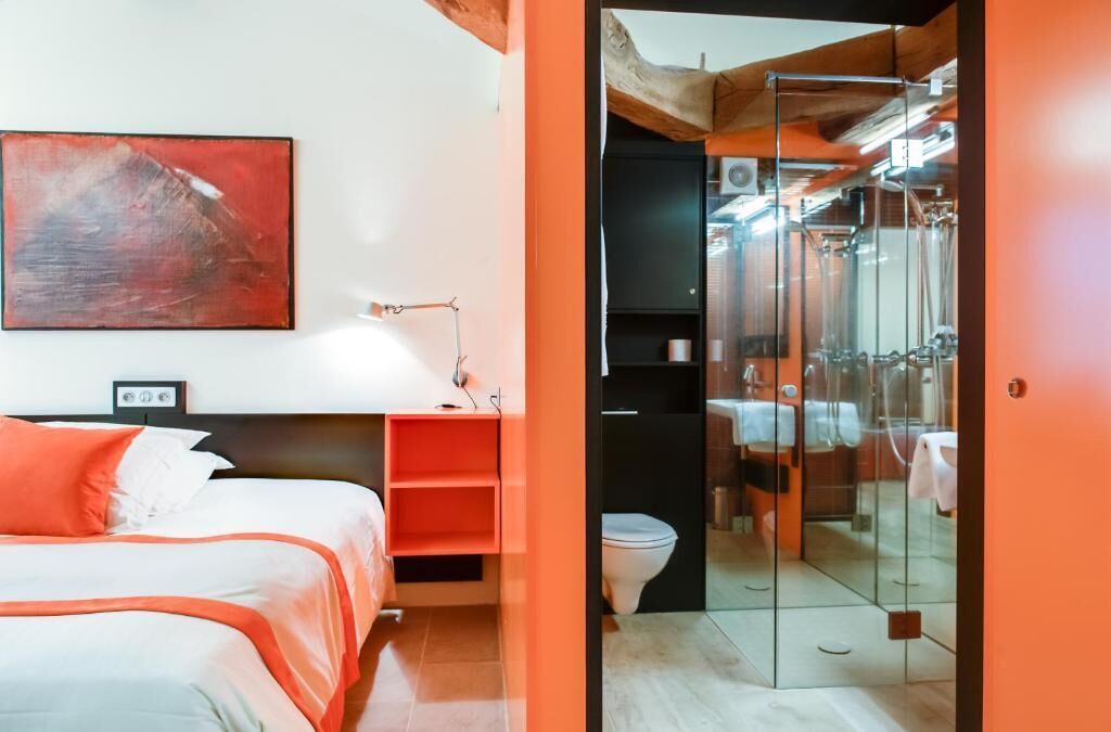 A modern hotel room with a bed, a painting, a desk, and a bathroom visible through a glass door.
