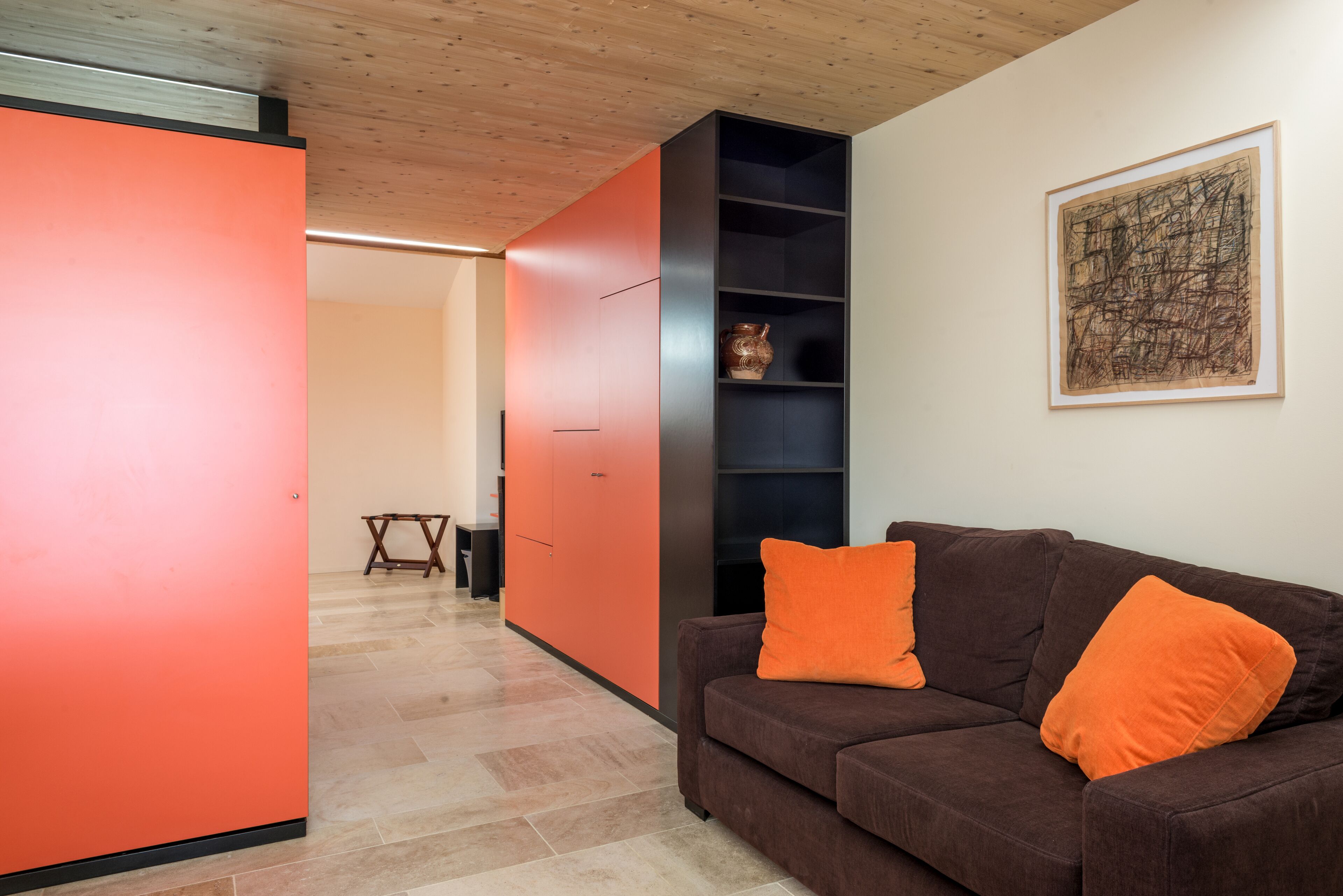 A modern living room with a brown sofa and orange cushions, a black shelving unit, and a framed abstract painting on the wall.