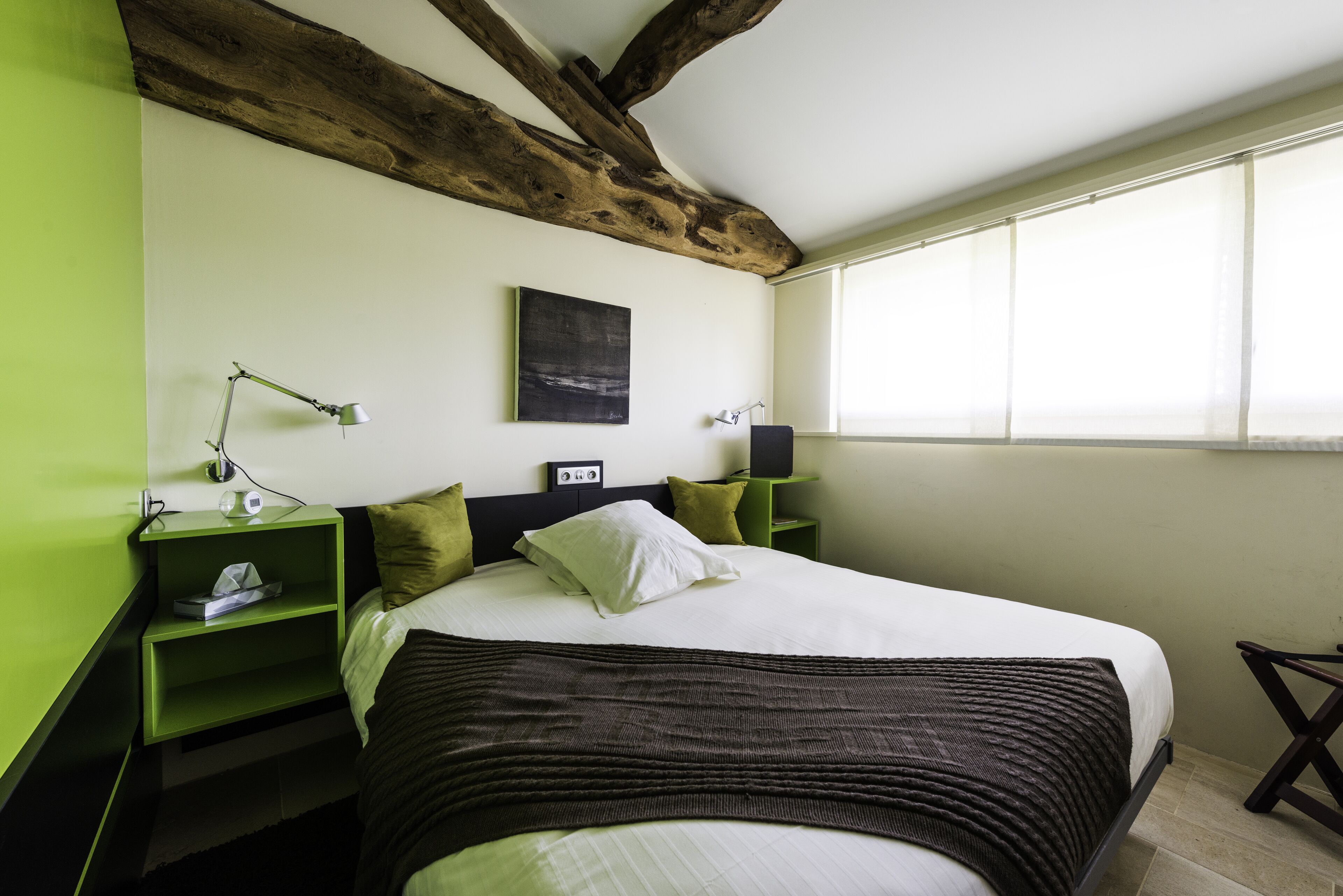 Une chambre avec un grand lit, des tables de chevet vertes, un plafond avec une poutre en bois, un tableau et une fenêtre.