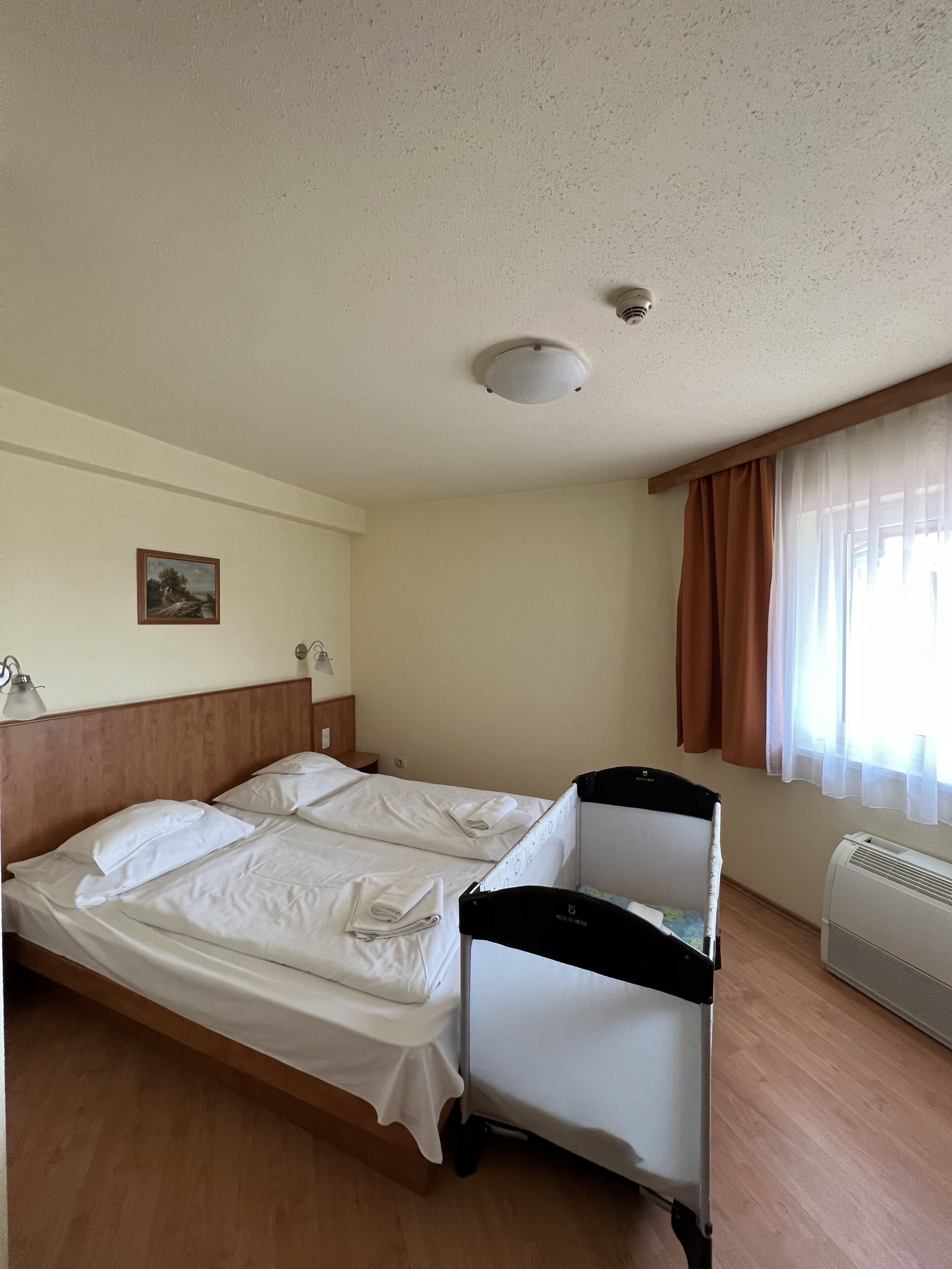 Une chambre d’hôtel avec deux lits, un berceau, un luminaire au plafond, un tableau et une fenêtre avec des rideaux.
