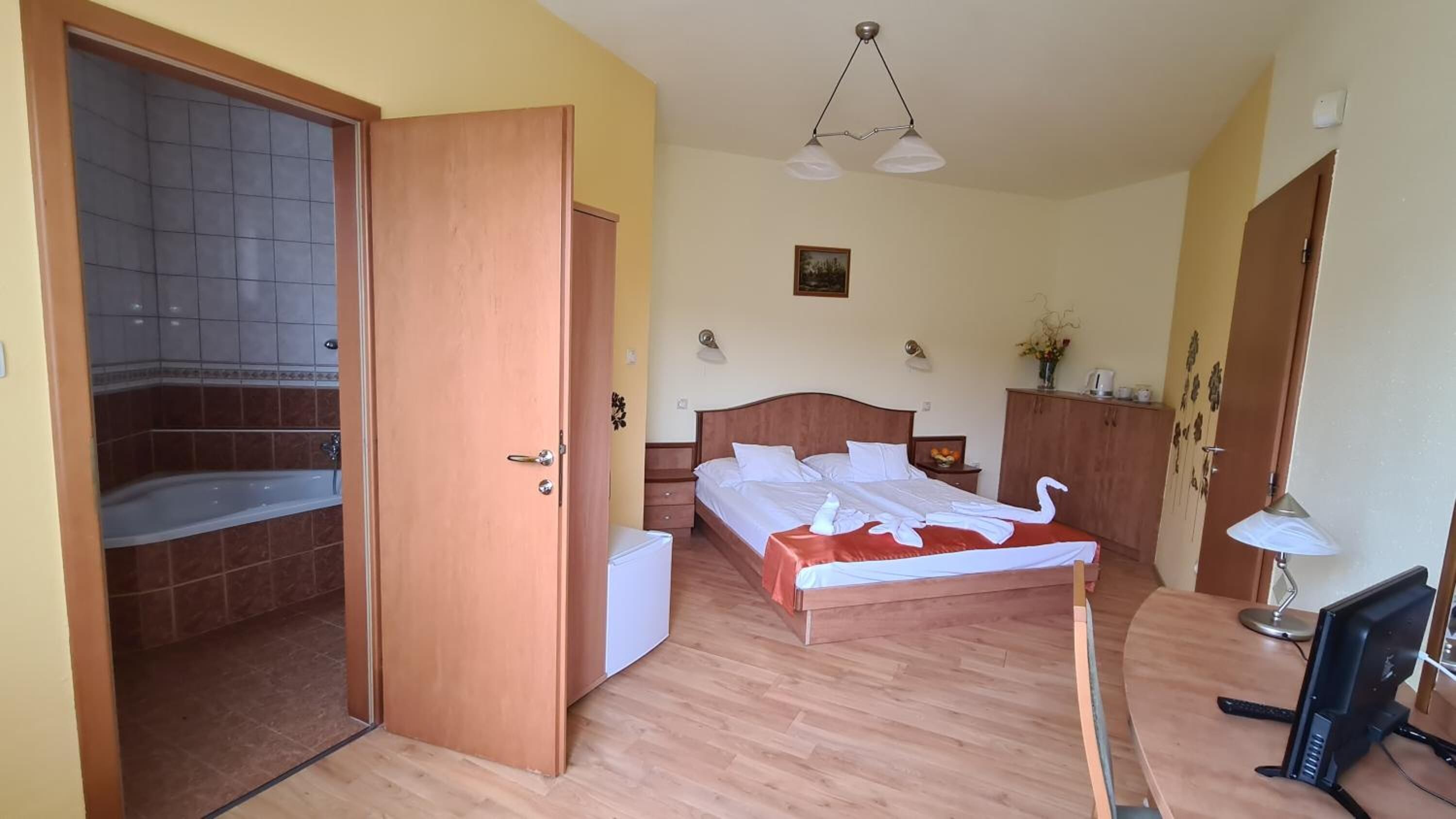 Una habitación de hotel con una cama de madera, un escritorio con computadora, una lámpara de escritorio, una mesita con un florero y un baño con bañera.