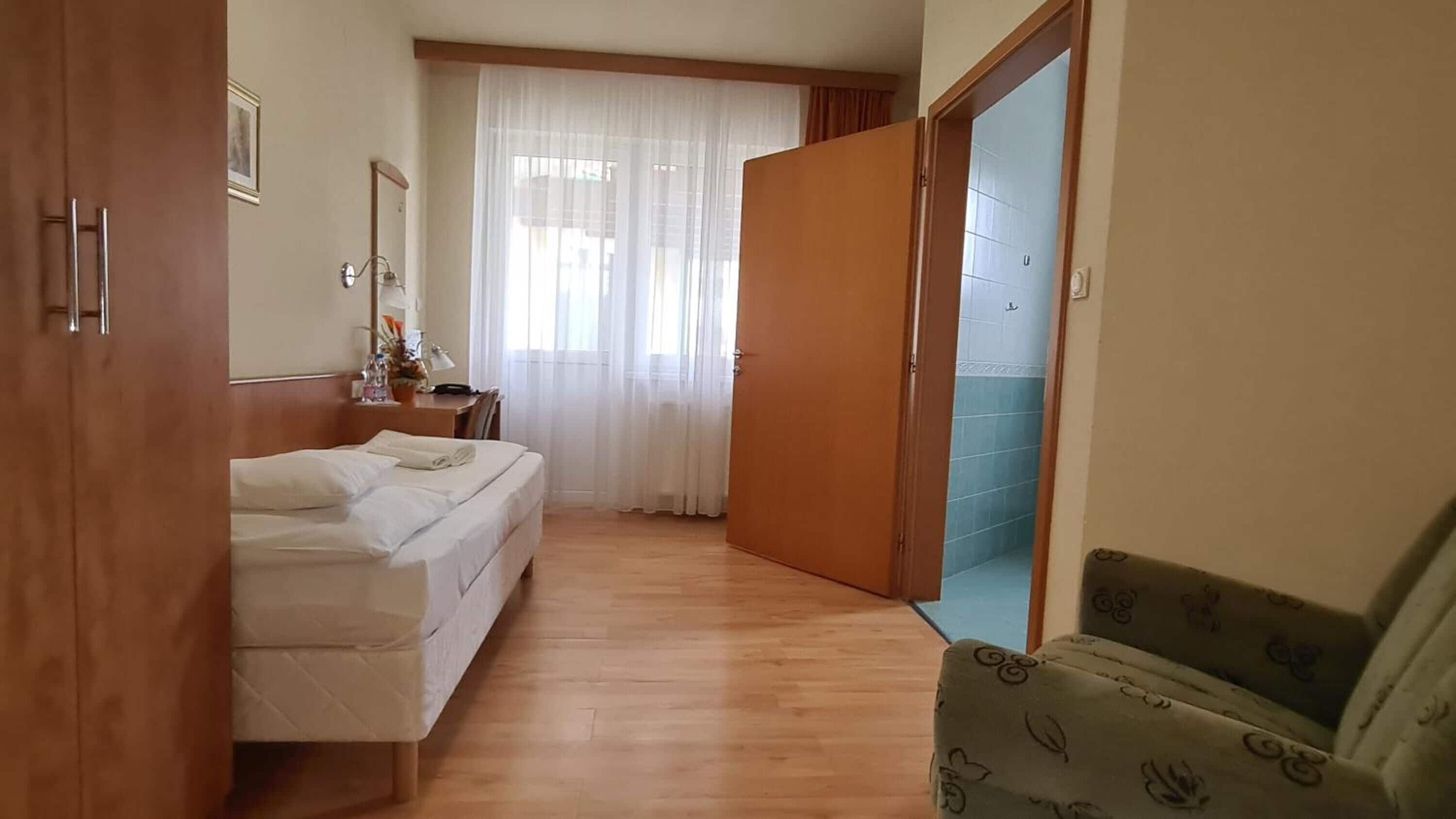 A hotel room with a bed, a desk with a chair, a mirror, and a bathroom visible through an open door.